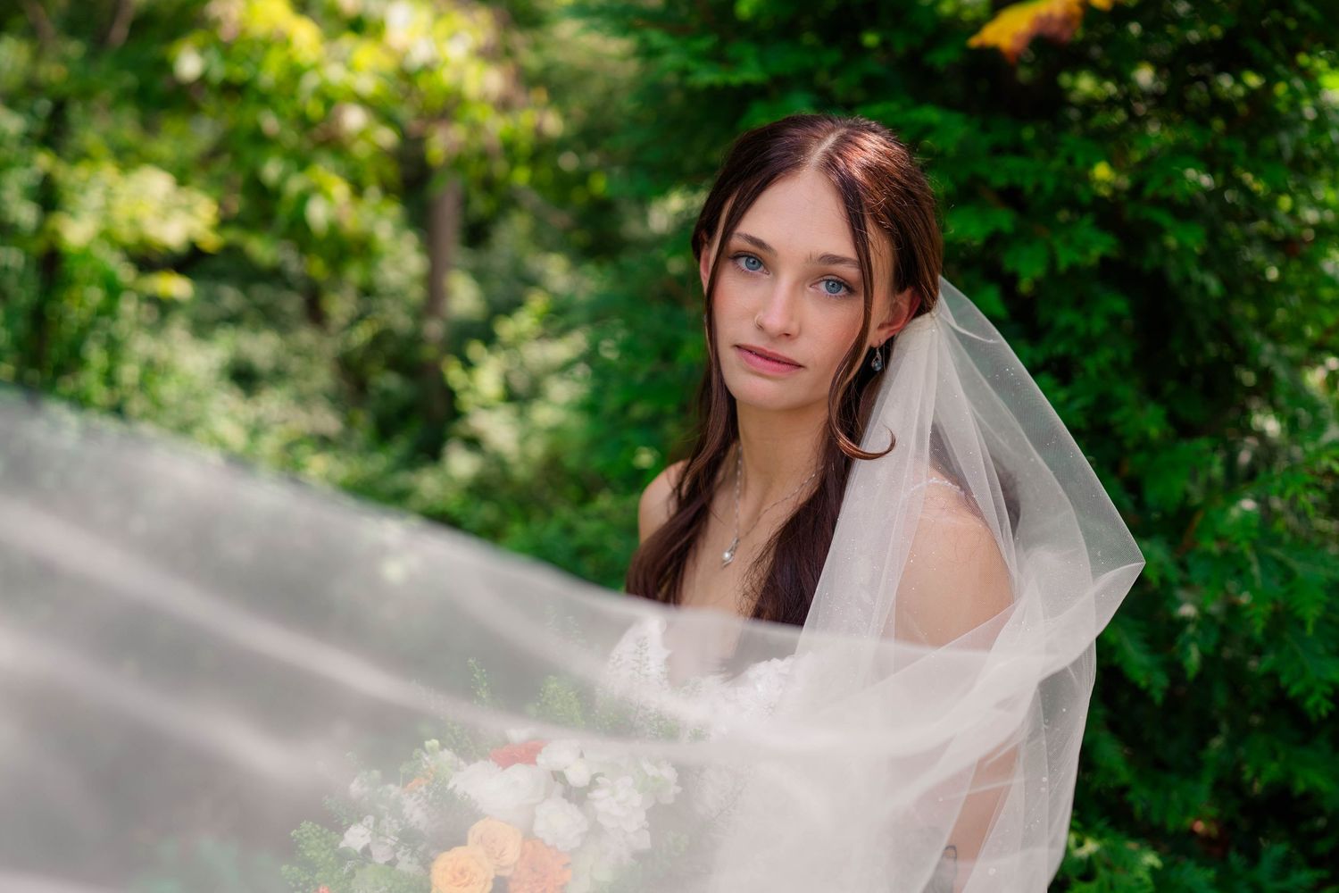 Elegant bride in white veil poses outdoors surrounded by lush green foliage holding bouquet.