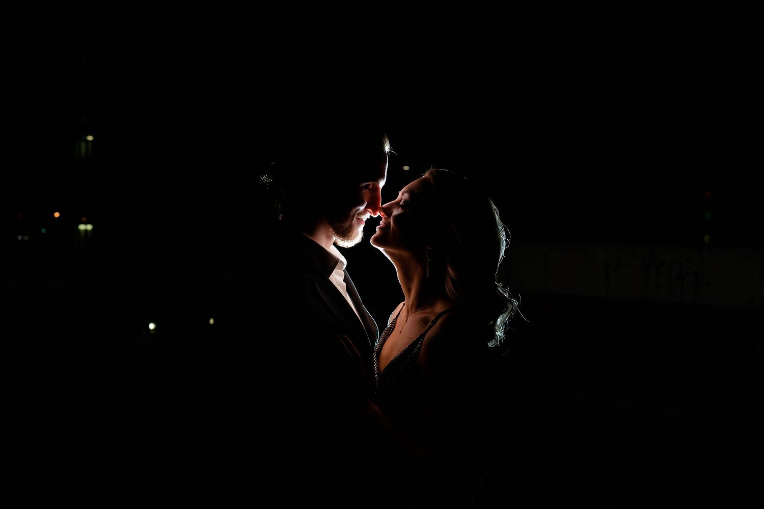 Intimate silhouette of a couple kissing in near darkness with faint city lights glowing softly behind them.