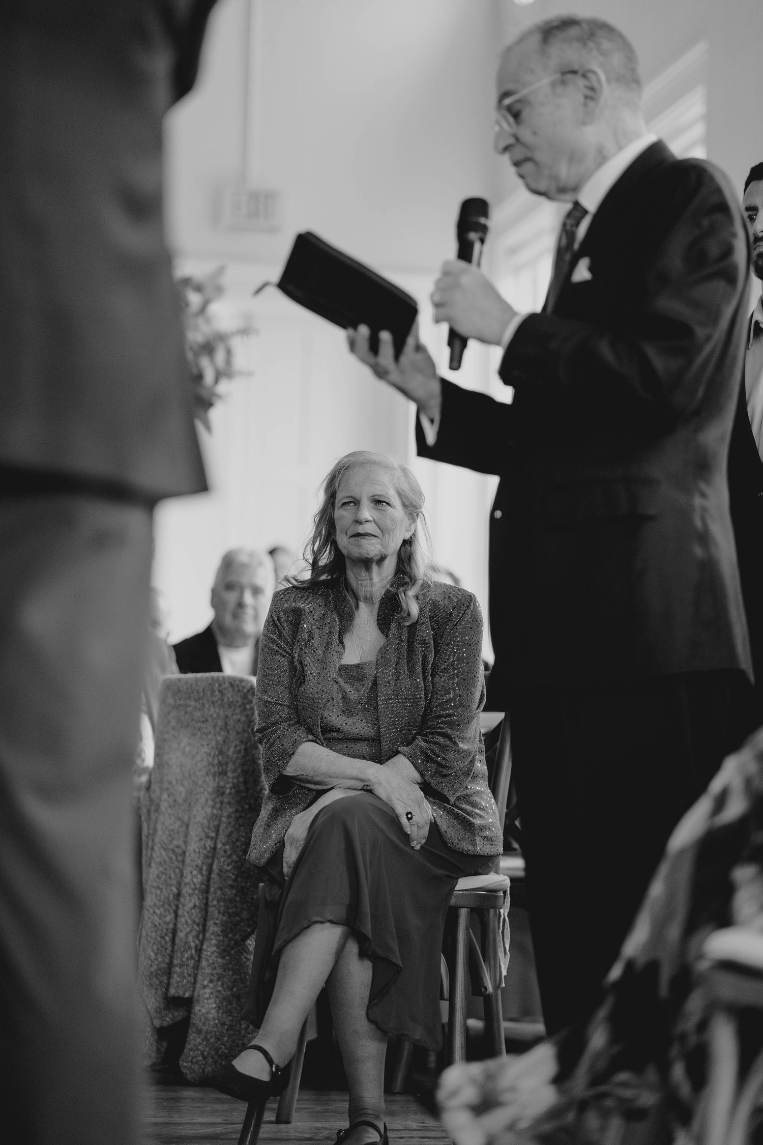 Black and white photo of elderly woman seated while man reads at wedding ceremony.