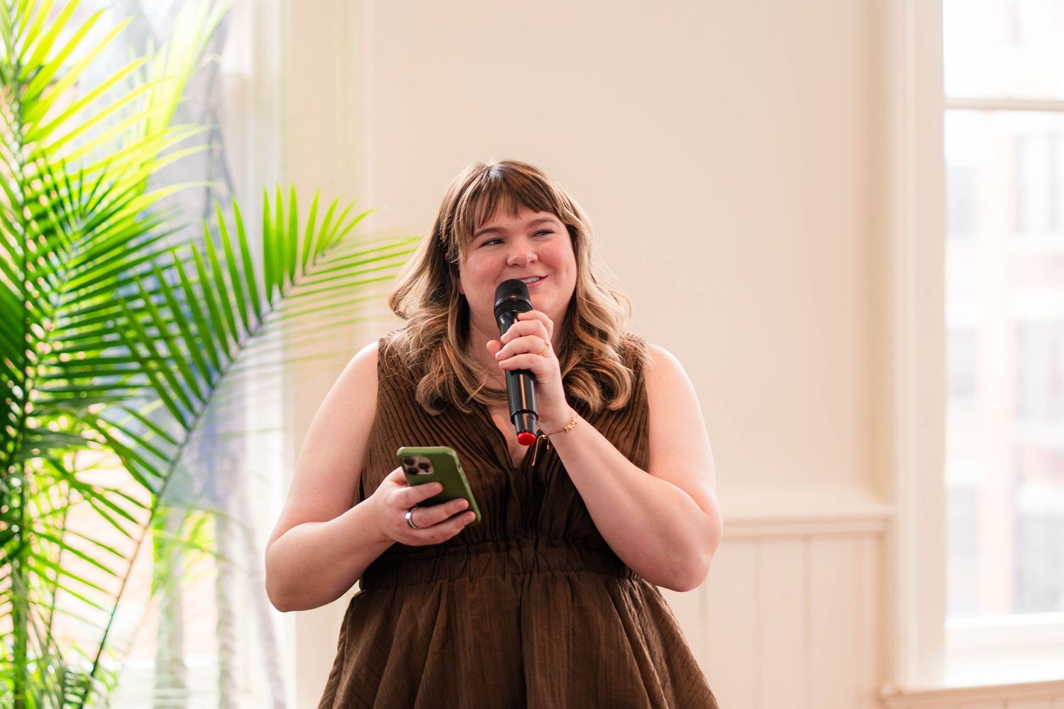 Woman in brown ruffled dress speaking into a microphone while holding a phone at a wedding reception.