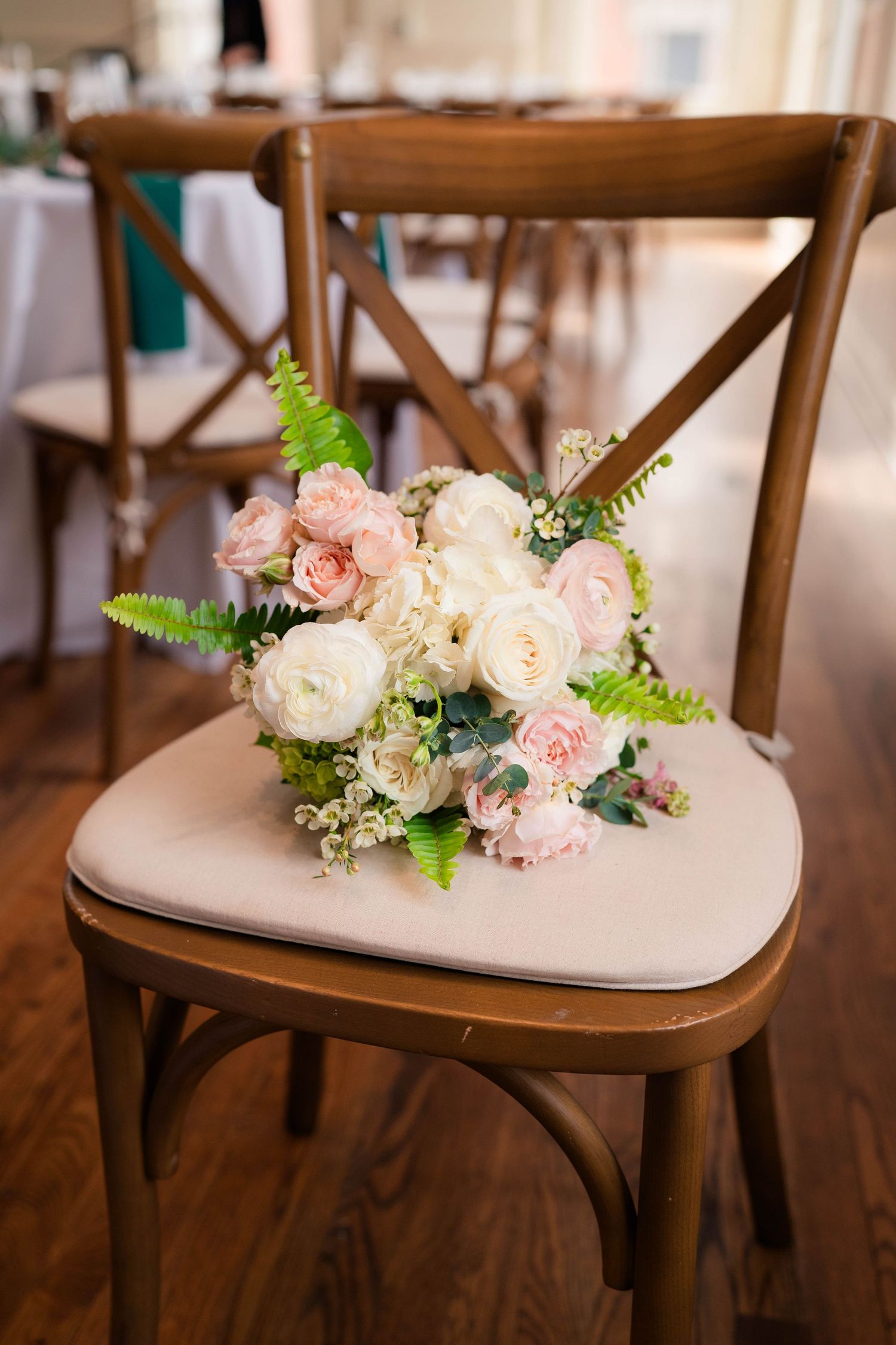 Bridal bouquet with blush and white roses resting on a wooden cross-back chair at a wedding reception.