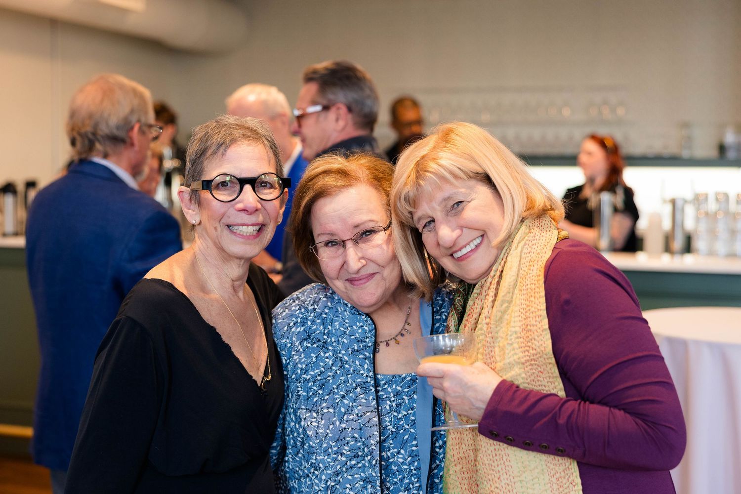 Three women smiling and posing together at a social gathering event.