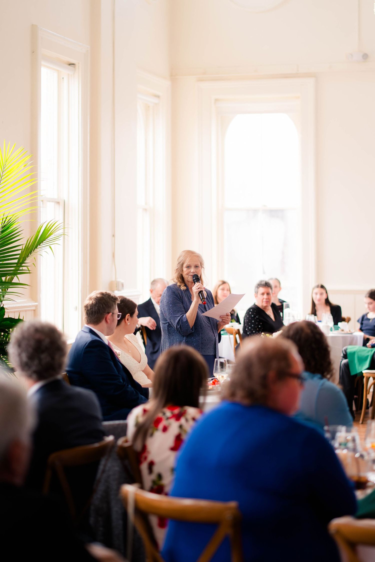 Wedding guest gives speech to seated reception guests in bright elegant venue.