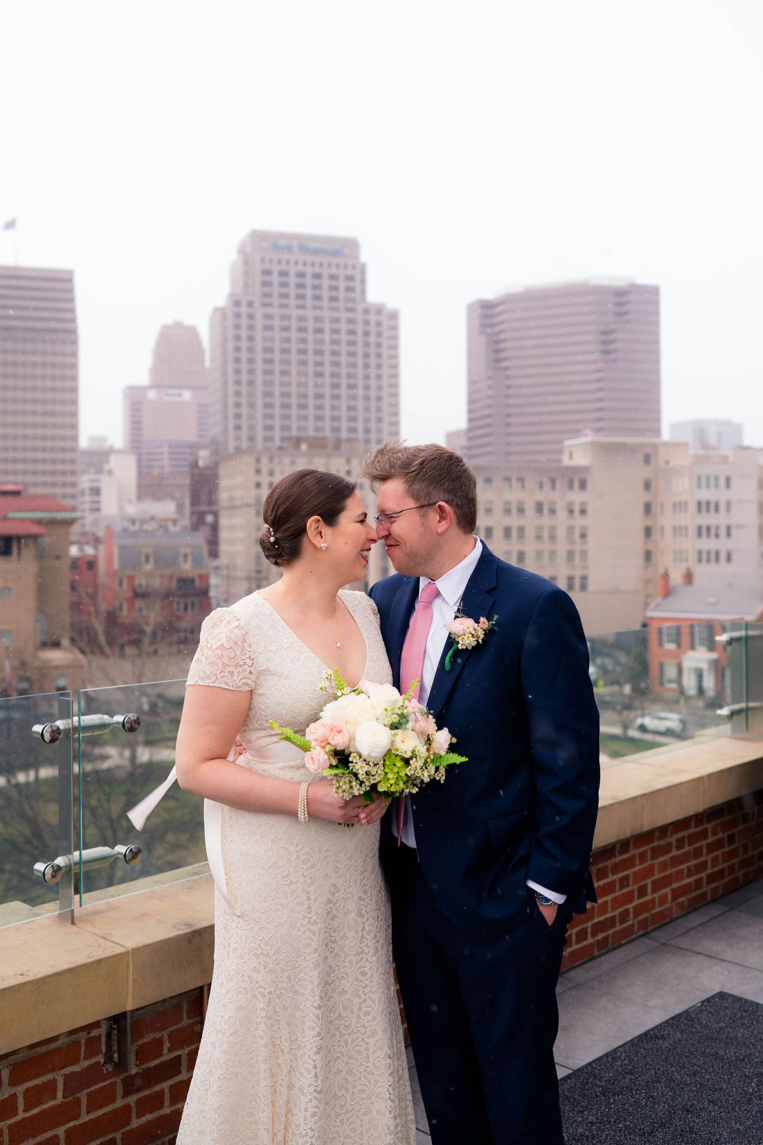Wedding couple touching foreheads tenderly on rooftop terrace with tall city buildings in background.