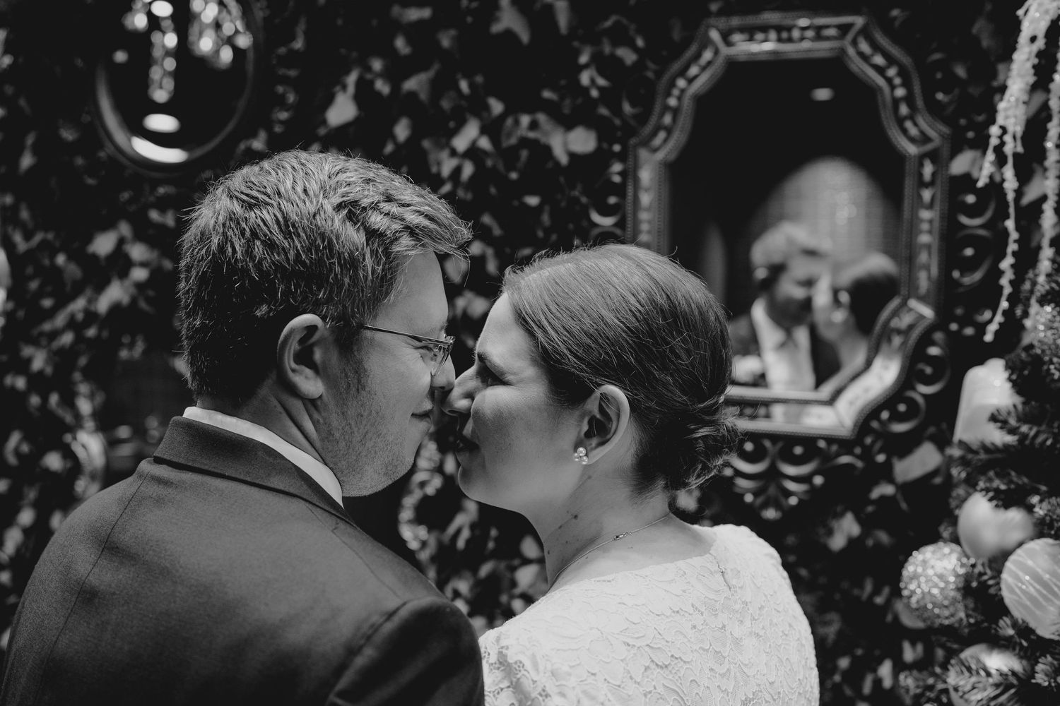 Black and white photo of bride and groom sharing an intimate kiss near a decorative mirror with holiday ornaments.