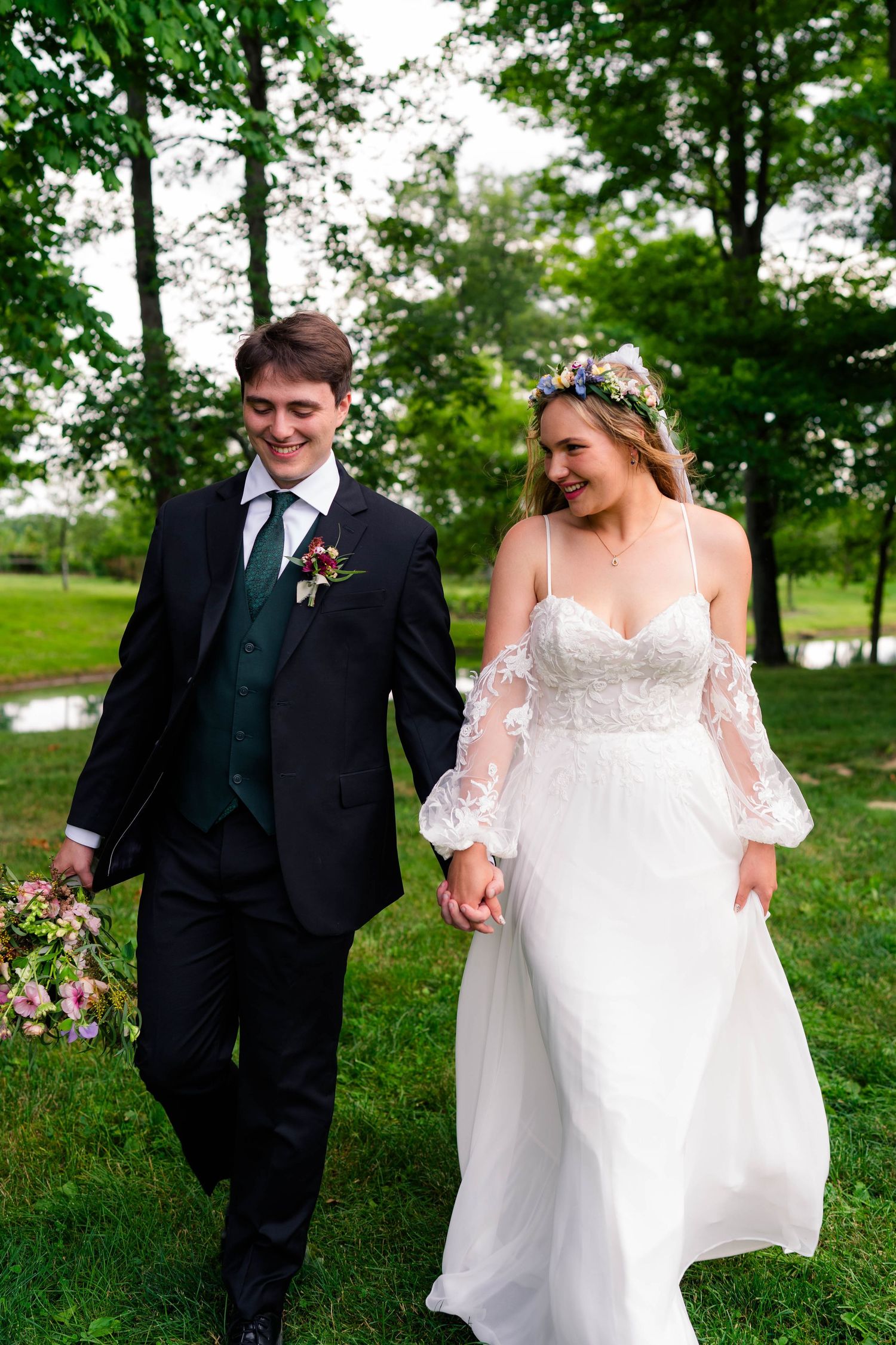 Smiling bride and groom walk hand-in-hand through green park on their wedding day.