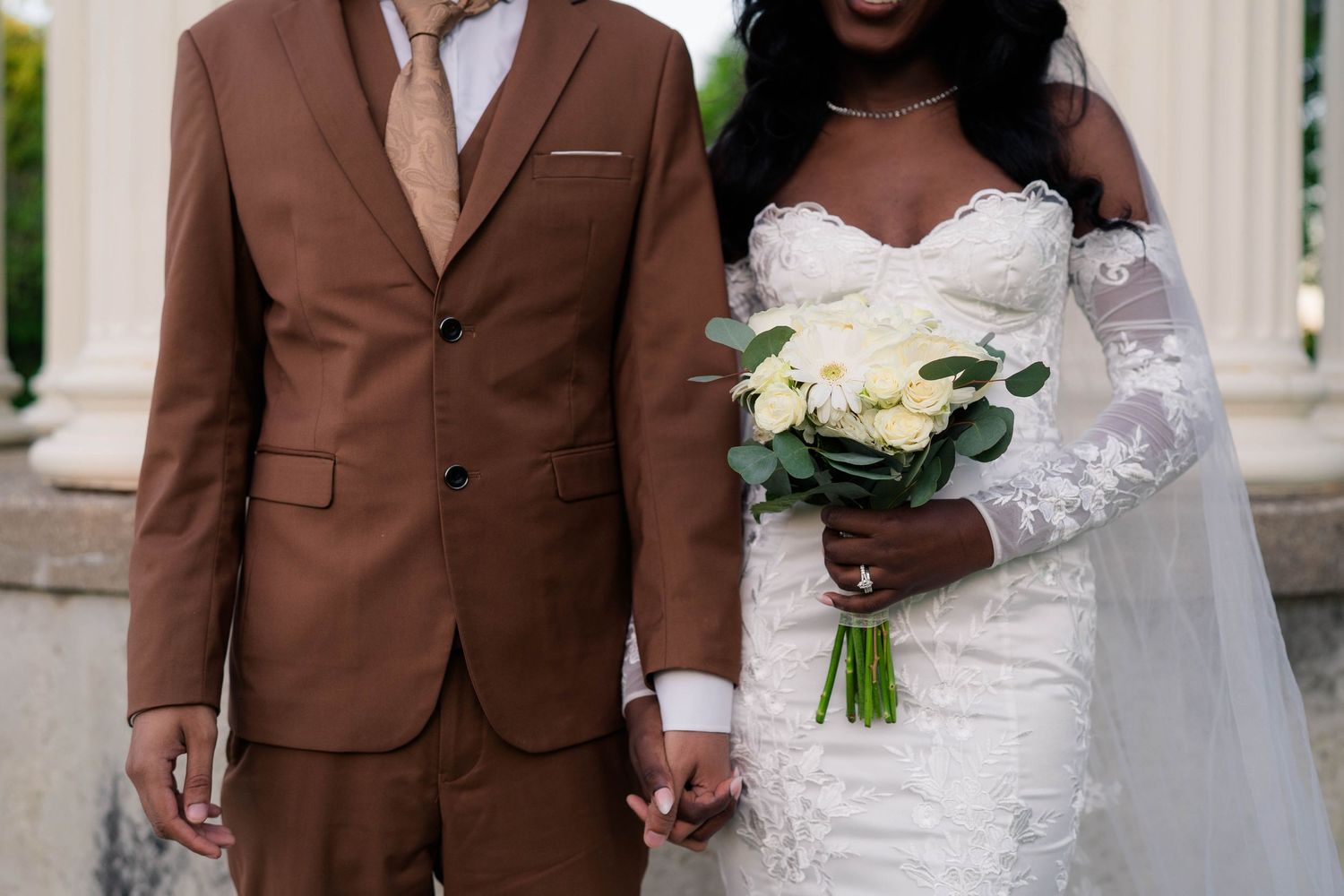 Couple in wedding attire hold hands, bride holds white bouquet in lace off-shoulder gown.