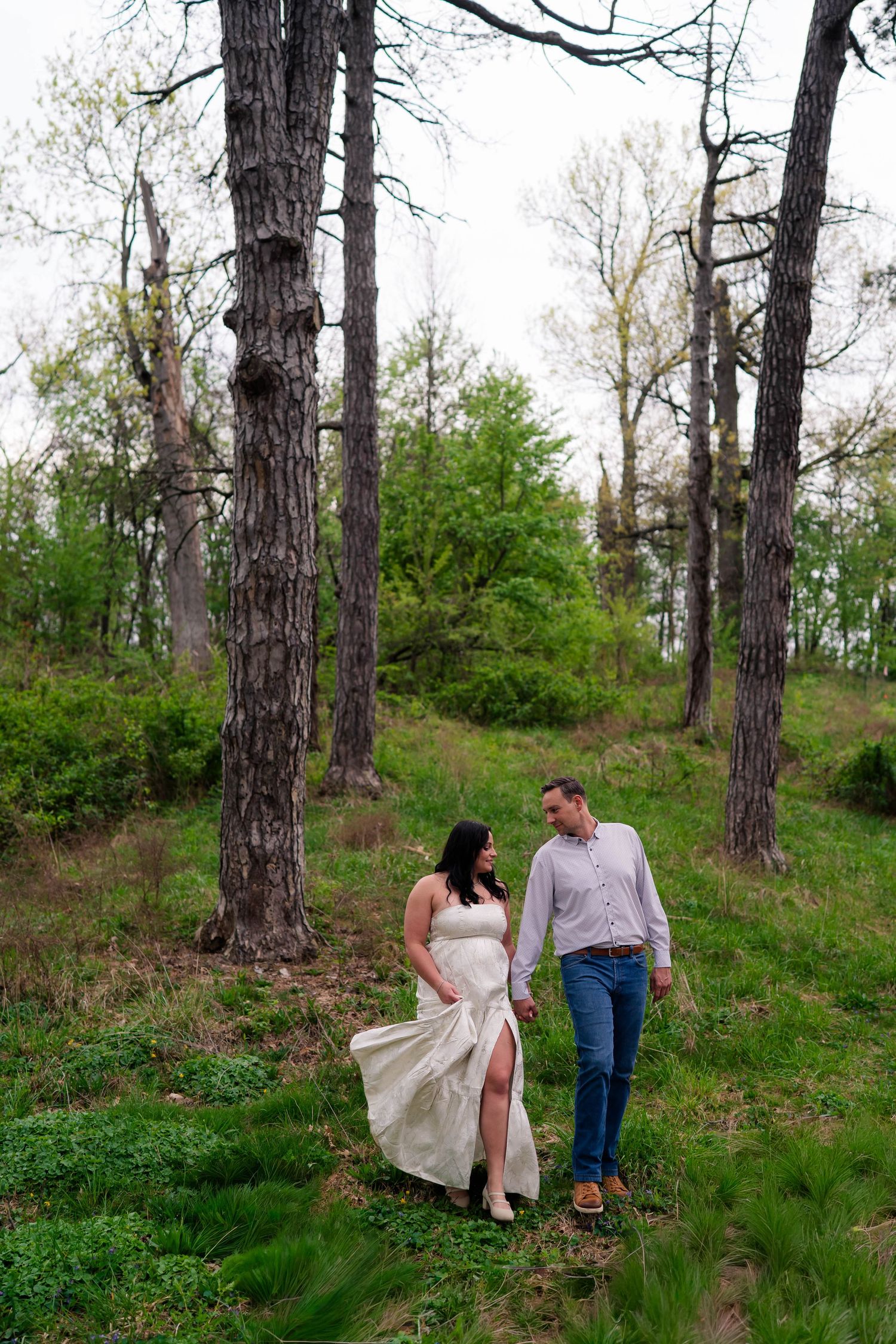 Couple walks hand-in-hand through lush green forest, woman in flowing white dress, man in blue jeans and lavender shirt.