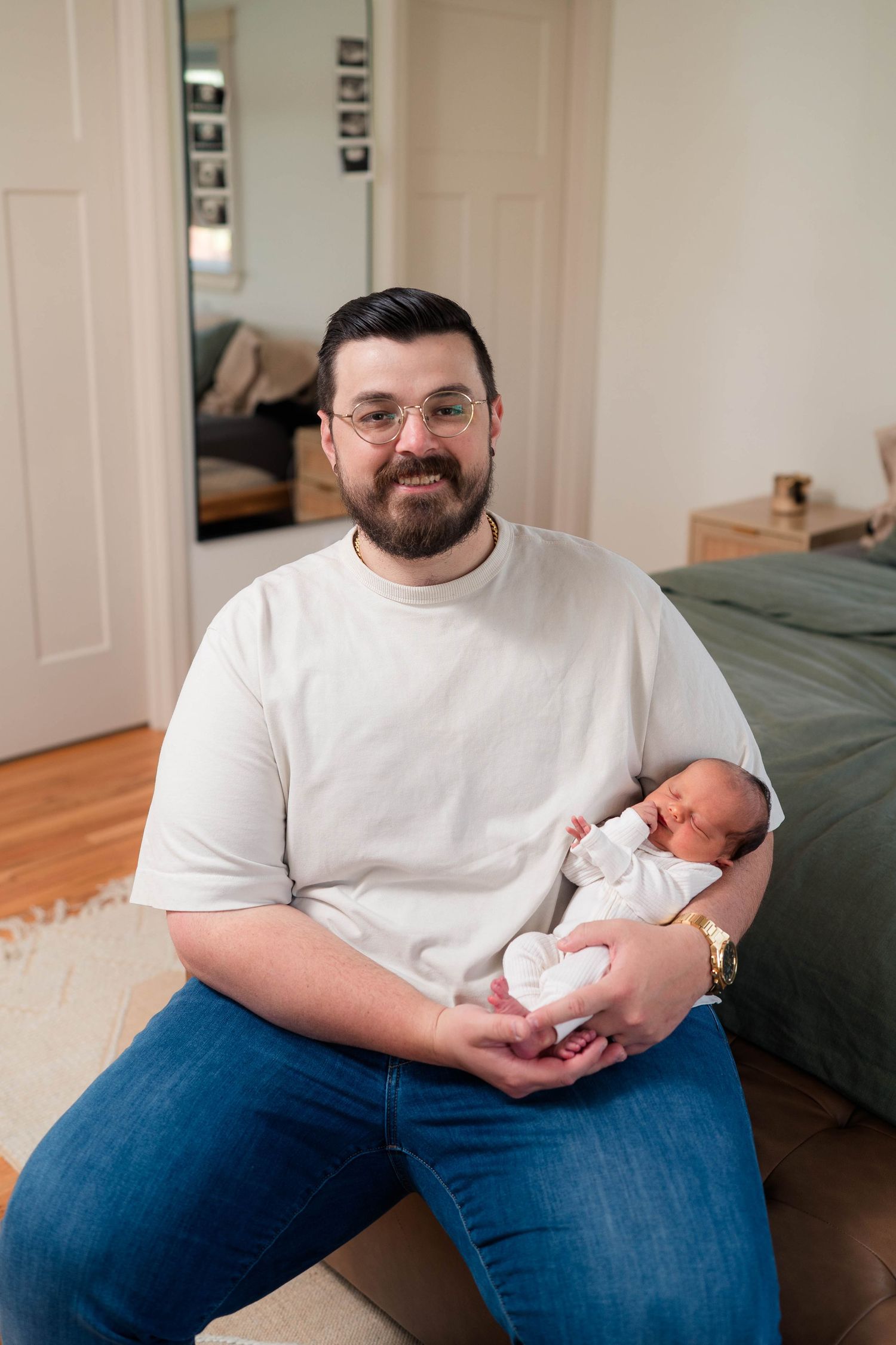 A bearded father wearing glasses and a white t-shirt sits smiling while cradling his newborn baby.