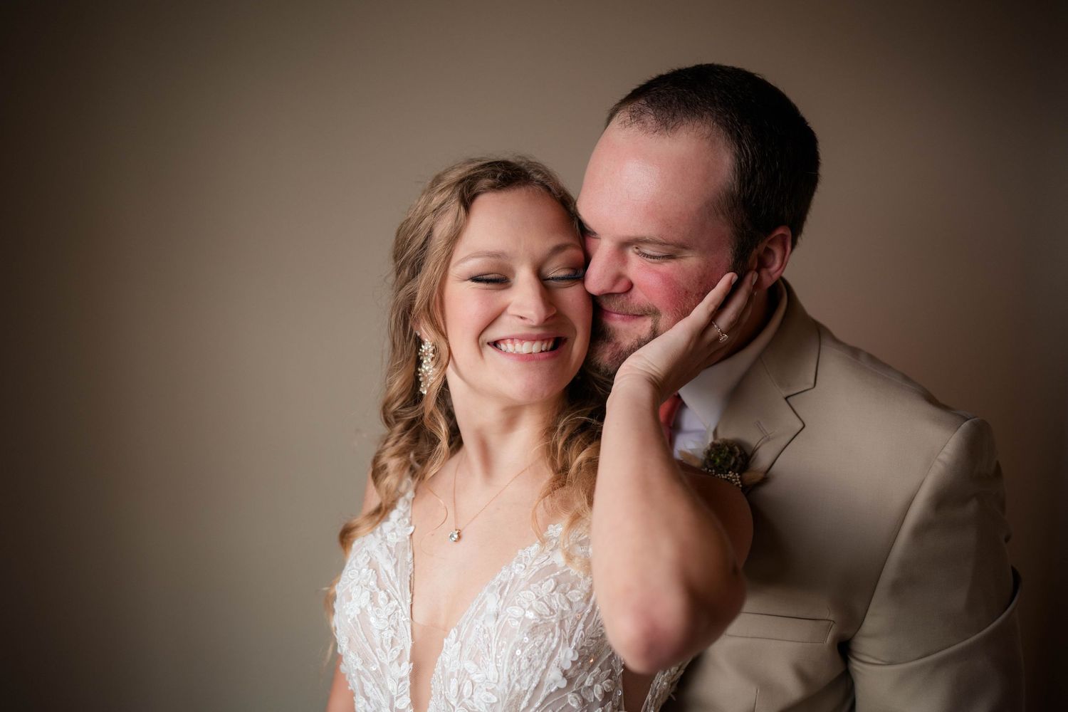 Groom tenderly kisses bride on cheek as she smiles joyfully in soft studio lighting.