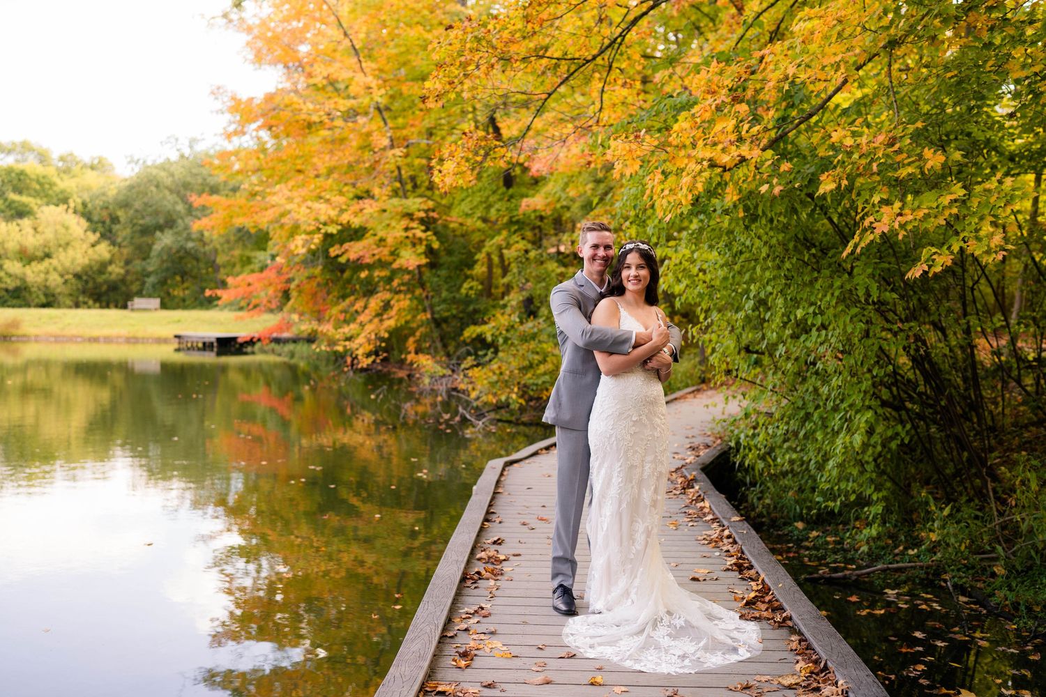 Bride and groom pose on wooden dock surrounded by vibrant autumn foliage reflecting in calm lake.