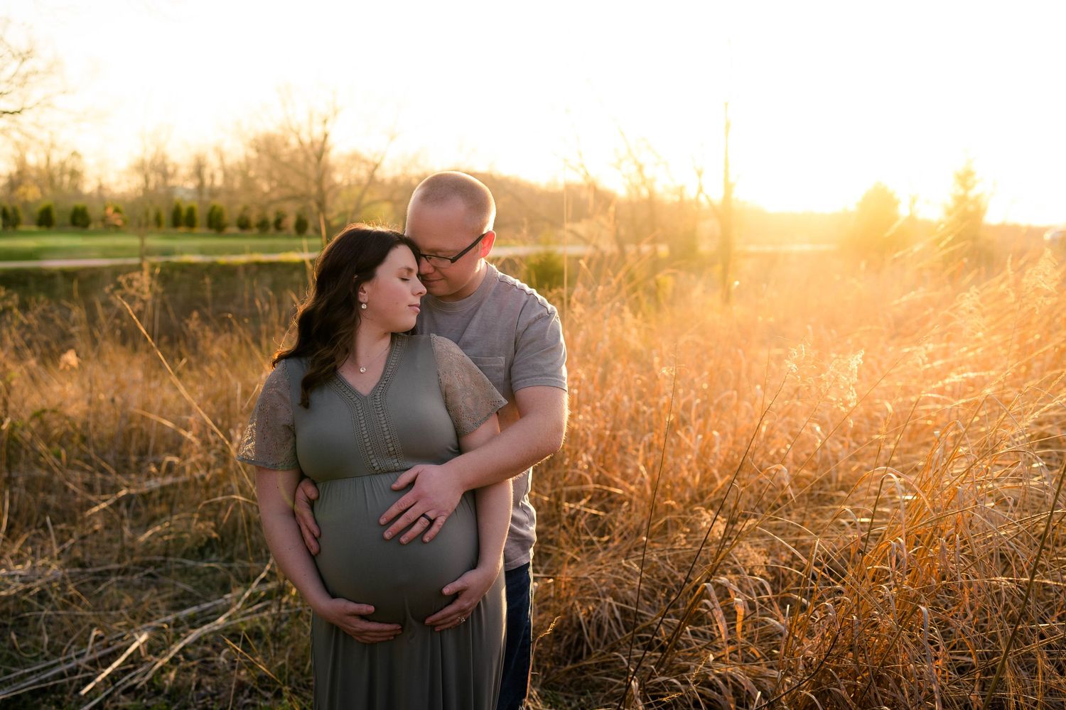 Couple sharing tender maternity photo at golden sunset in tall grass field, hands cradling baby bump.