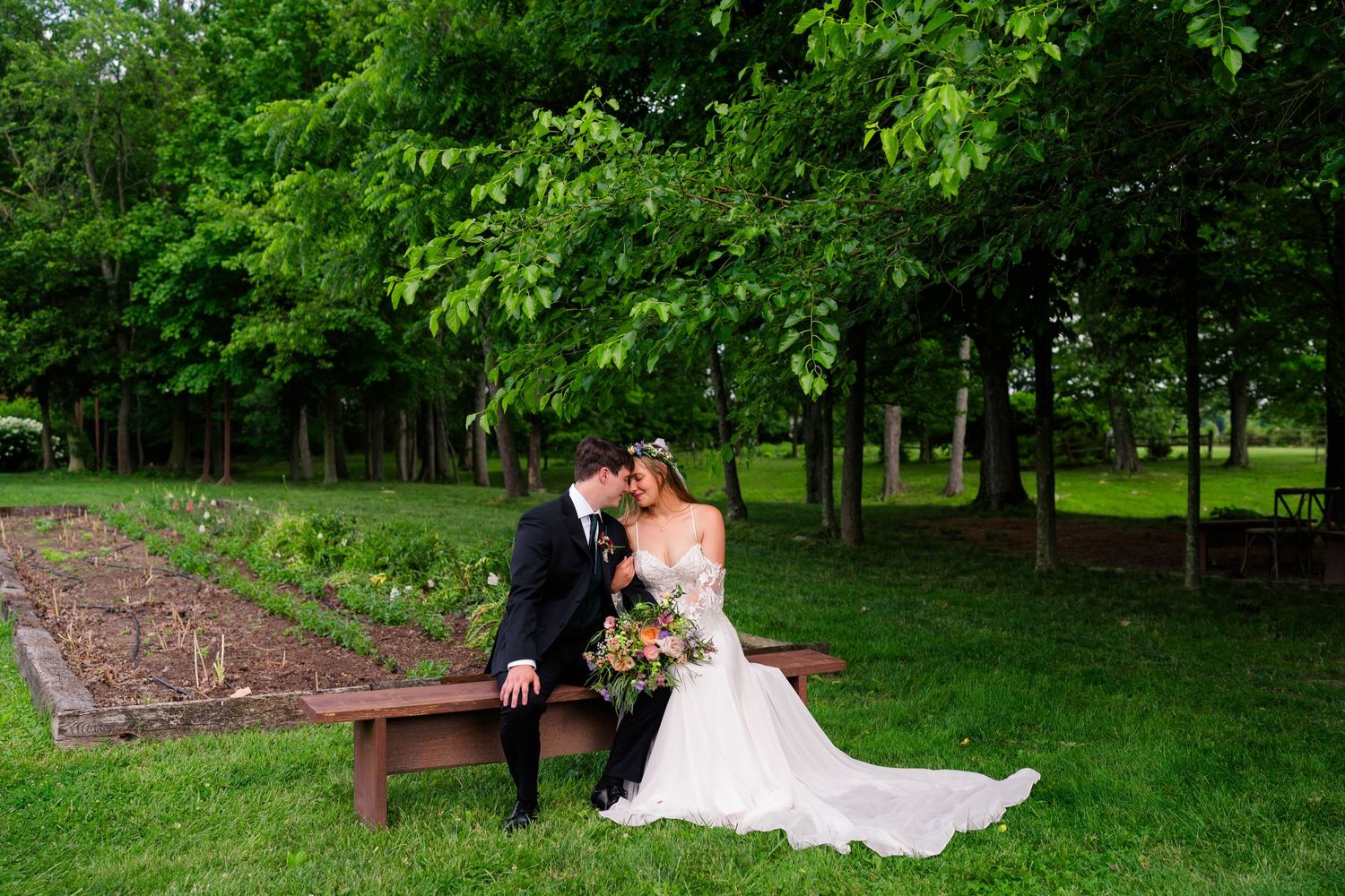 Bride and groom share intimate moment seated on wooden bench surrounded by lush green trees.