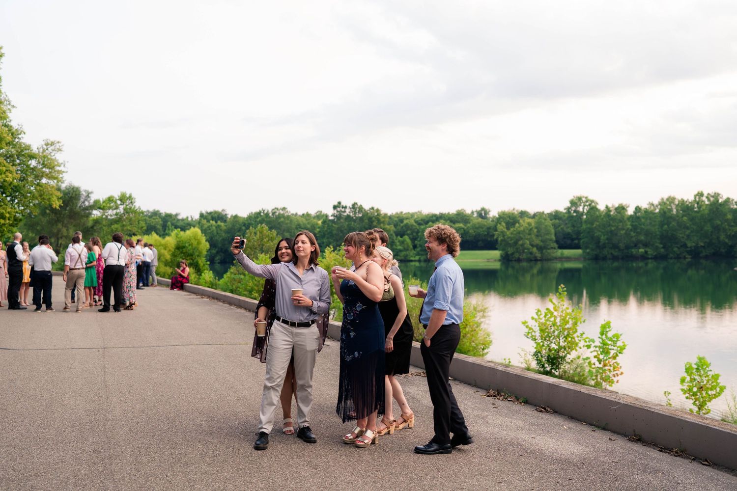 Wedding guests take selfie on lakeside promenade with scenic water views at sunset.