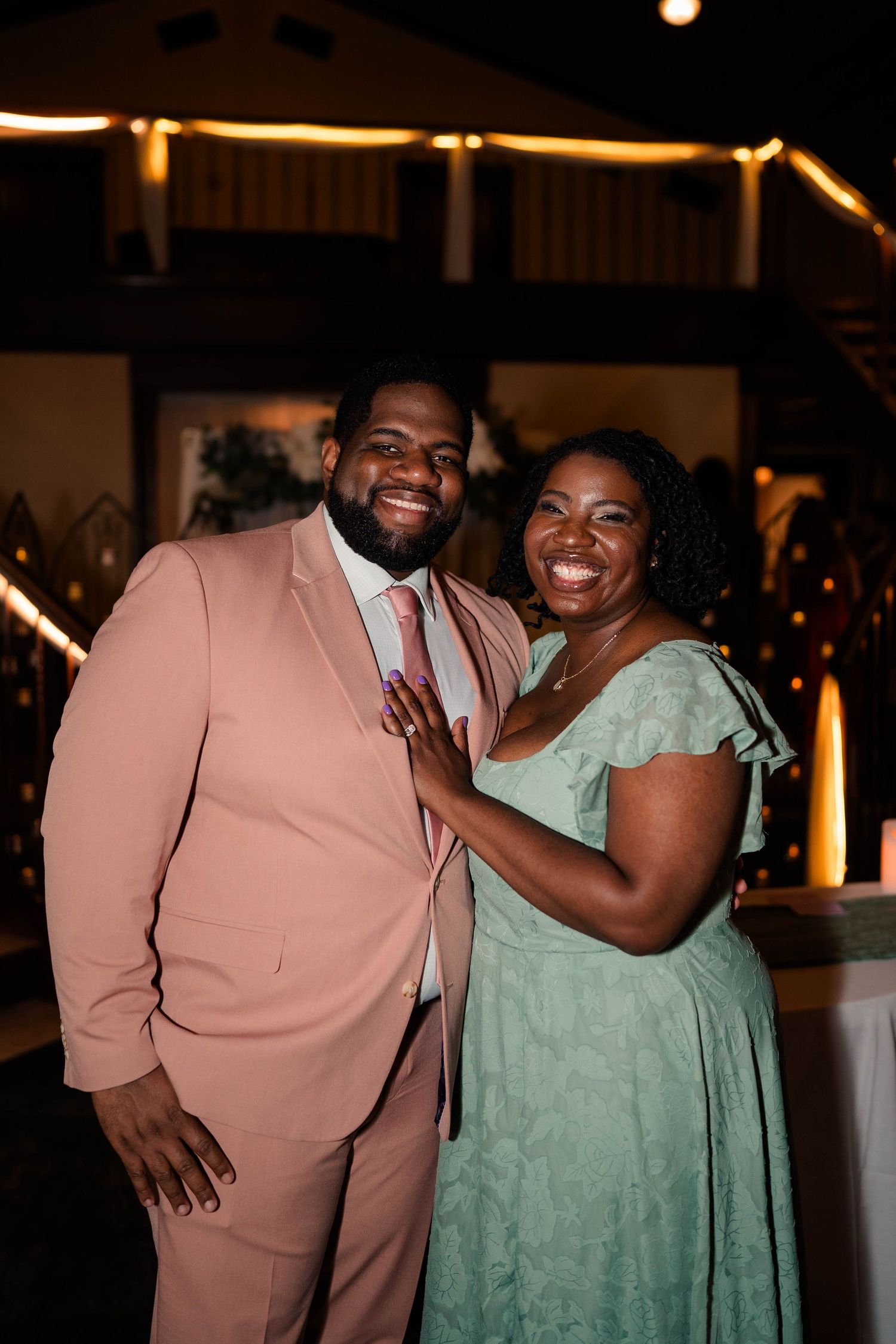 Couple smiling together at a wedding reception, man in pink suit, woman in green dress.