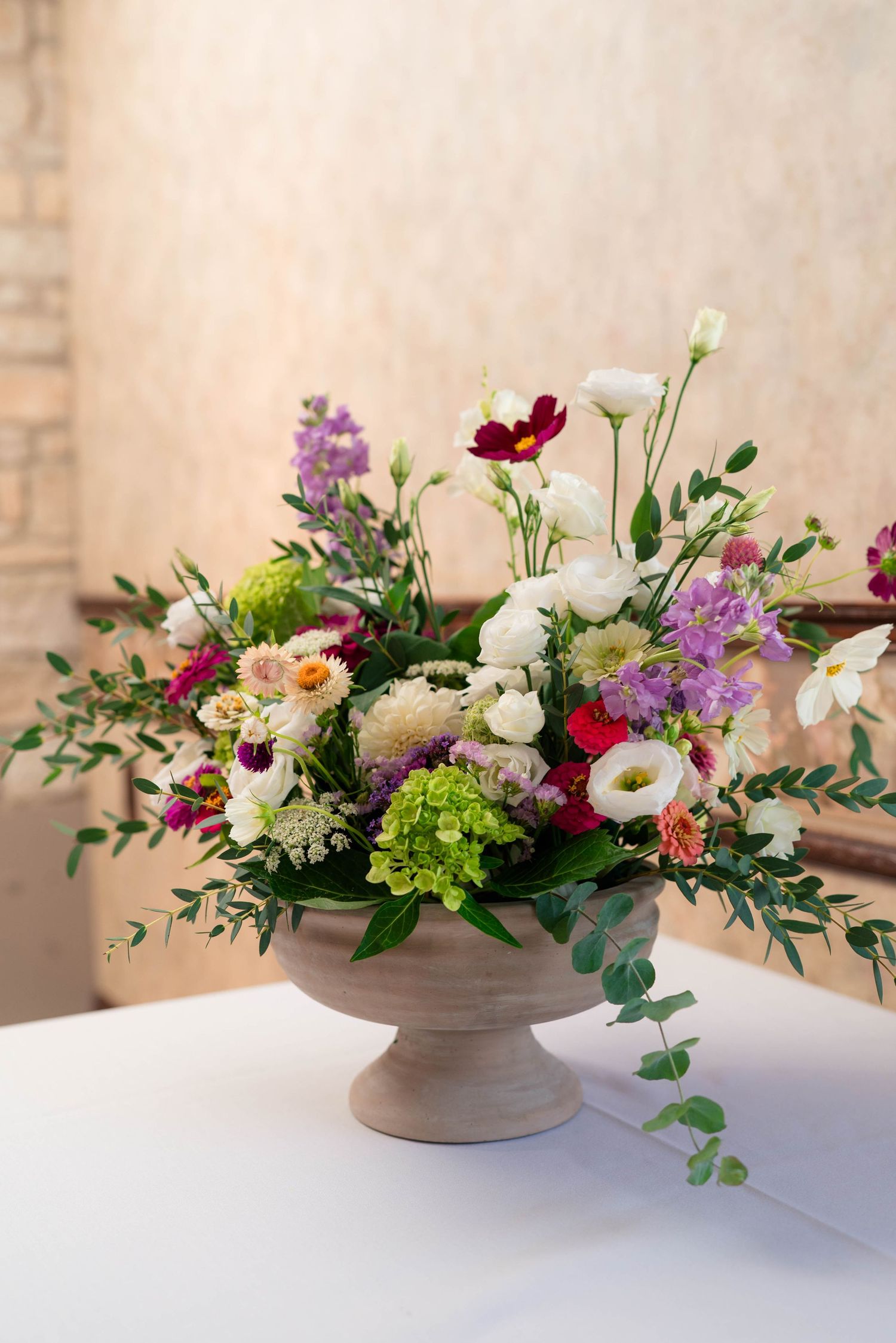 Elegant wedding centerpiece with colorful flowers in rustic pedestal vase on white tablecloth.