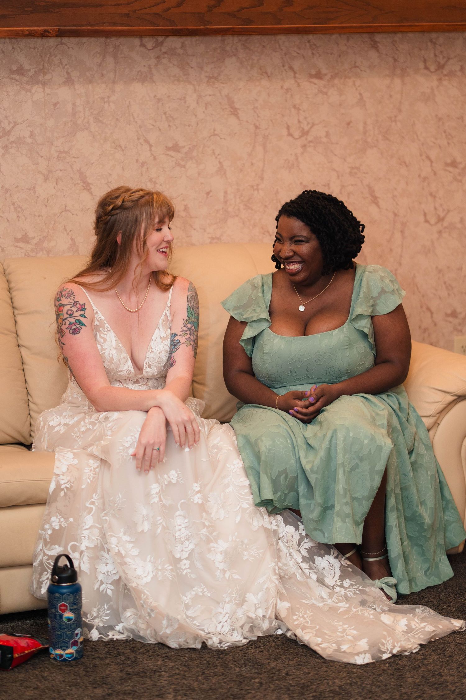 A bride in a lace wedding gown laughs with a bridesmaid in sage green on a cream sofa.