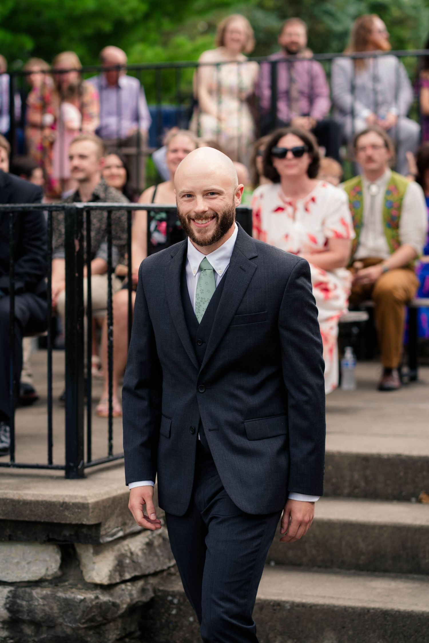 Smiling bald groom with beard walks down outdoor wedding ceremony steps wearing navy suit with sage green tie.