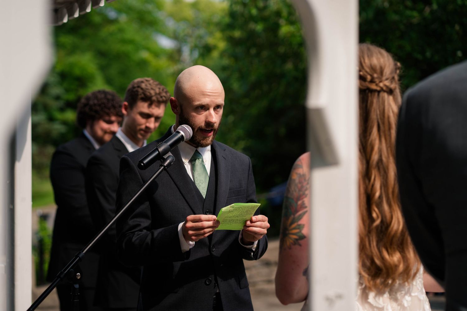 Bald officiant reads vows from green card at microphone during outdoor wedding ceremony with groomsmen standing behind.