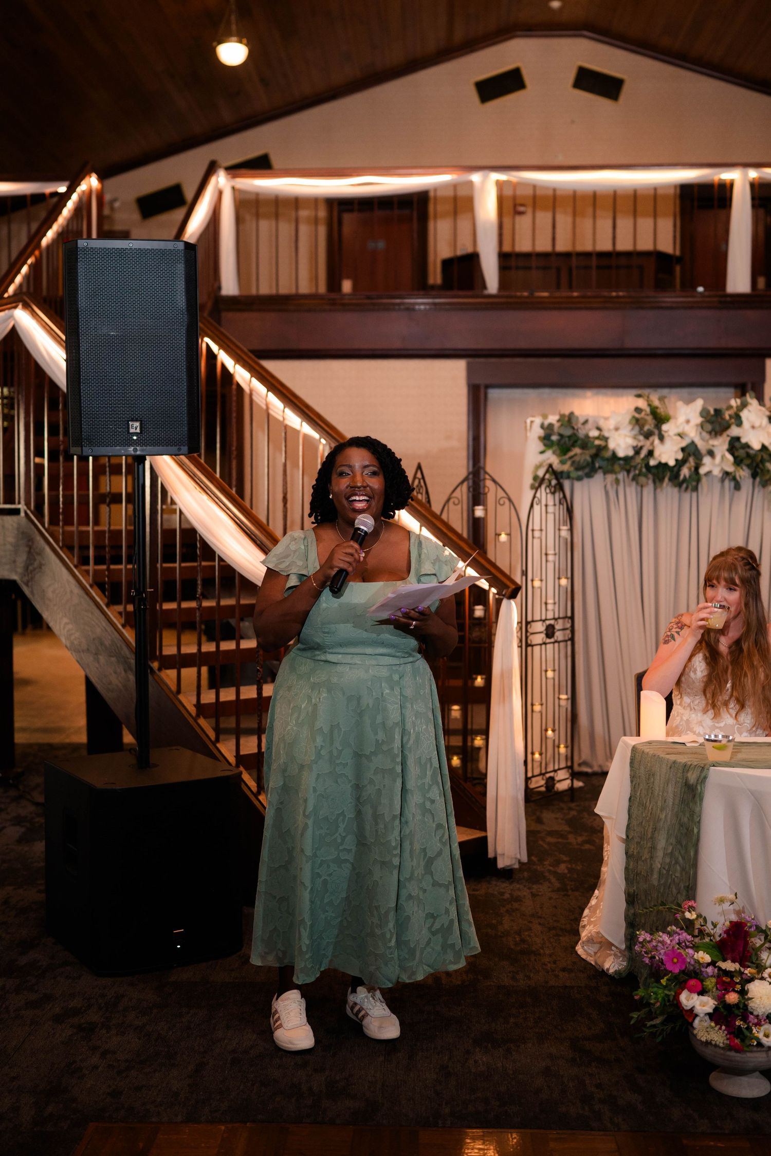 Woman in teal dress giving a speech at a wedding reception with microphone.