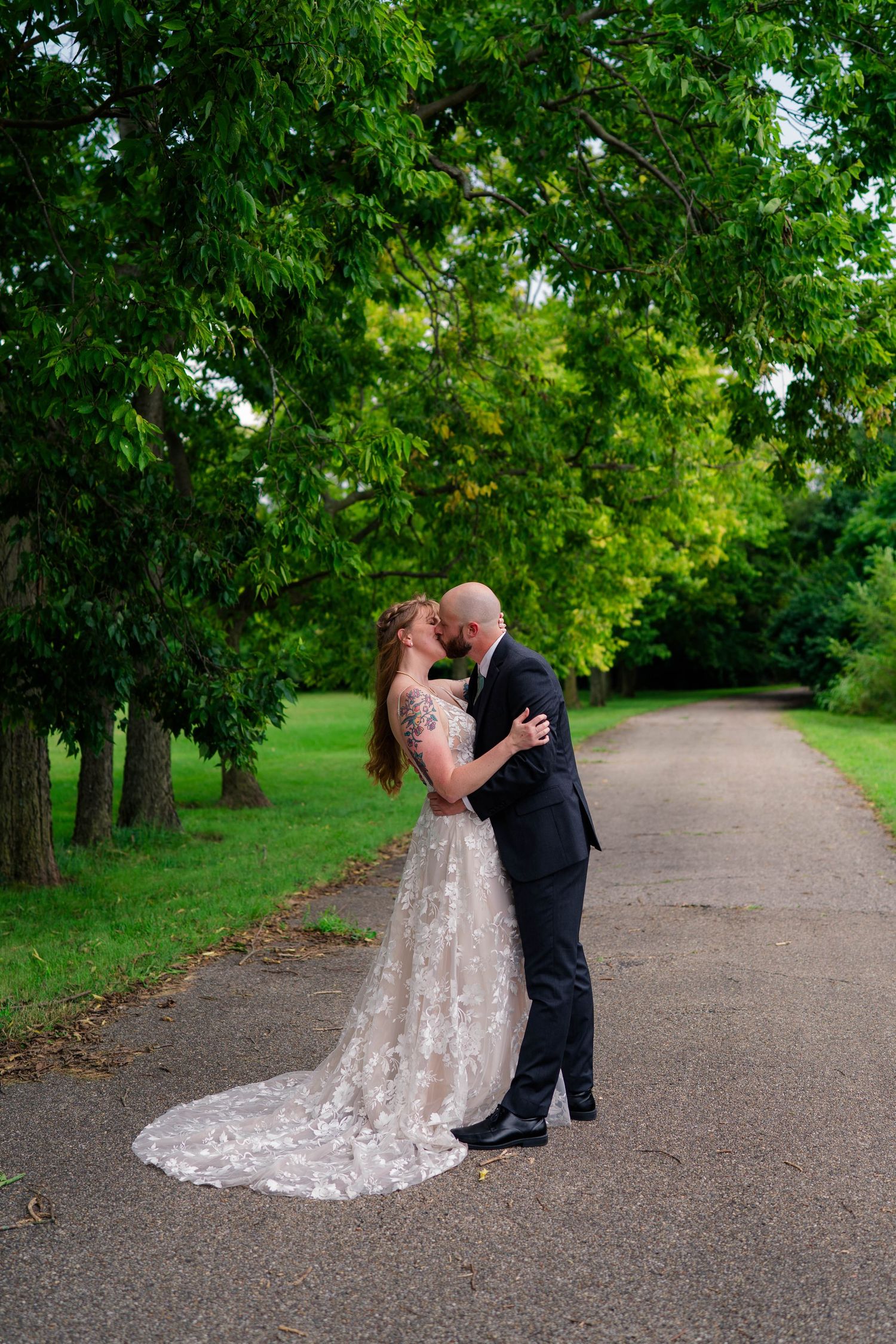 Bride and groom kiss on a tree-lined path, bride in lace gown with long train.