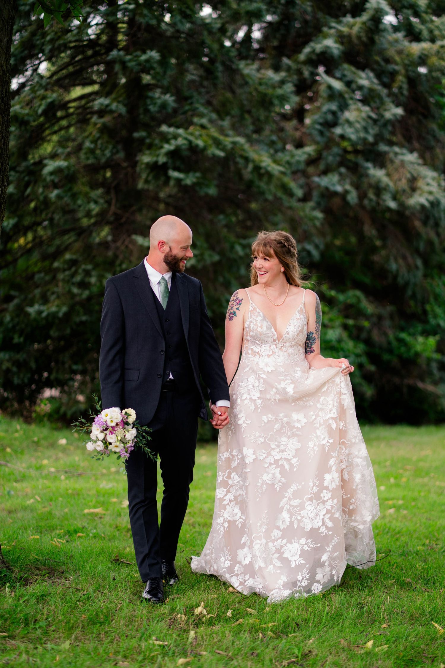 Bride and groom walk hand-in-hand through lush green park, bride wearing floral lace gown, groom in navy suit holding bouquet.