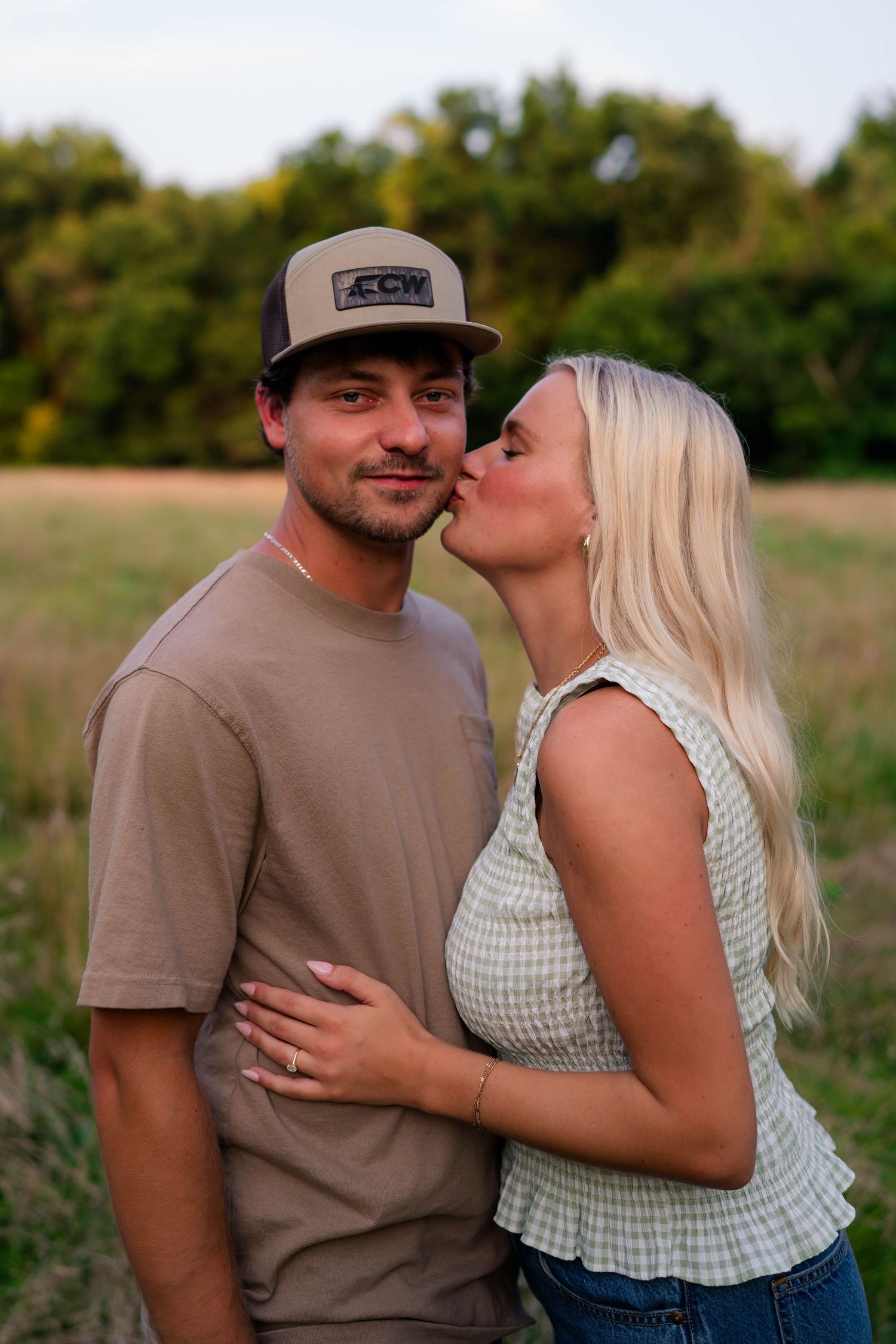 Blonde woman kissing man in brown cap and t-shirt on cheek during outdoor engagement shoot.