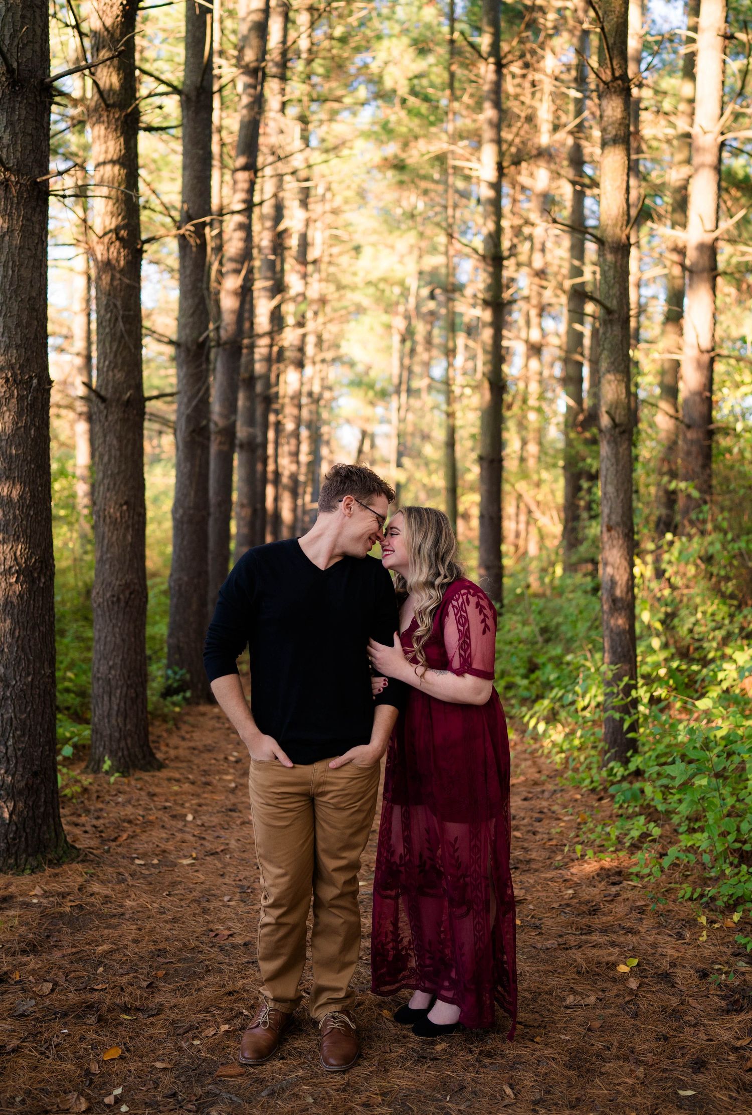 Couple sharing a gentle kiss on a sunlit forest path surrounded by tall pine trees.