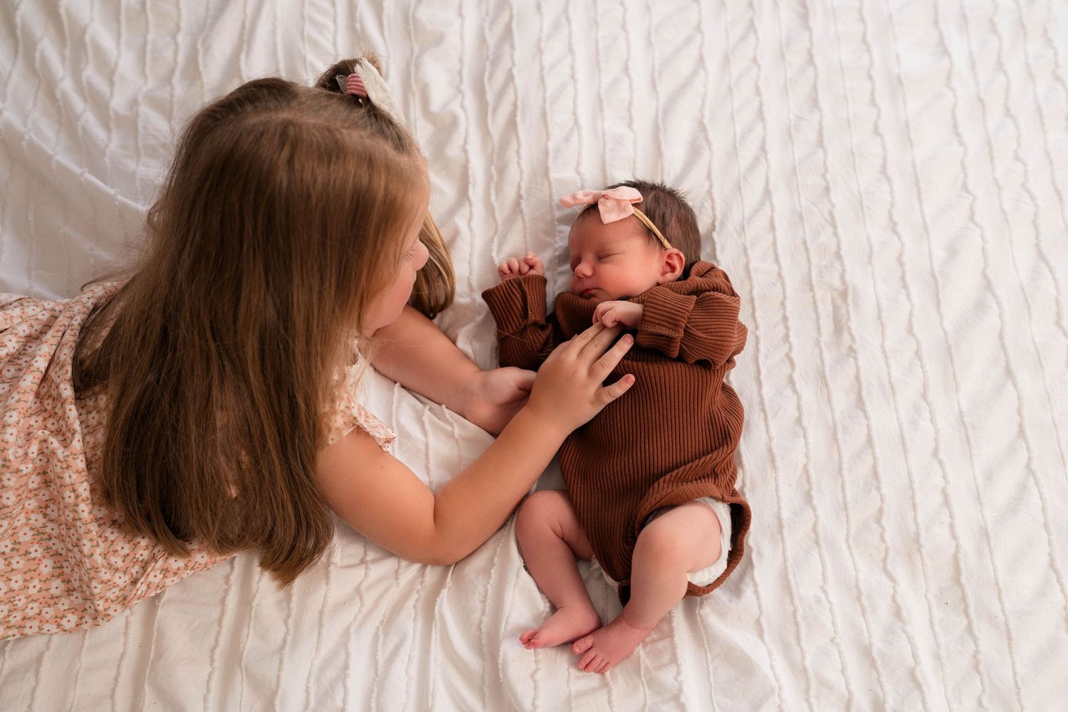 Young girl gently touches newborn baby sibling lying on white textured blanket, wearing brown outfit.