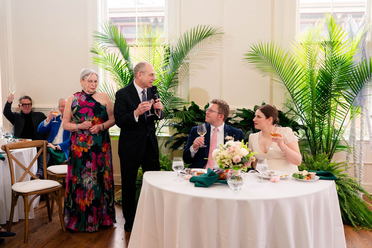 Parents giving toast at wedding reception as bride and groom raise glasses at head table.