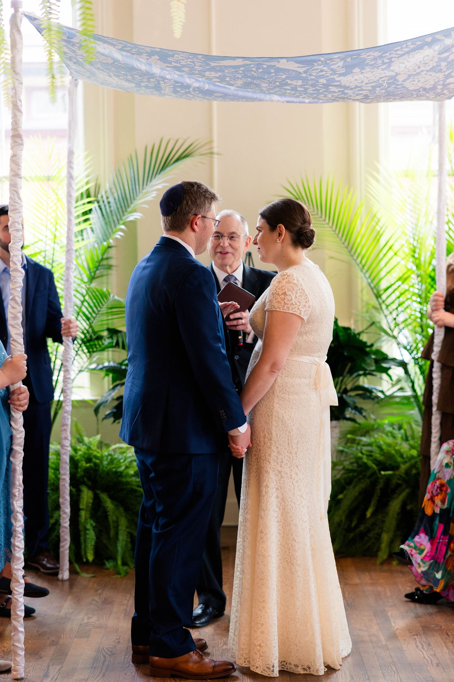 Bride and groom hold hands facing each other under a chuppah with palm leaves during indoor wedding ceremony.