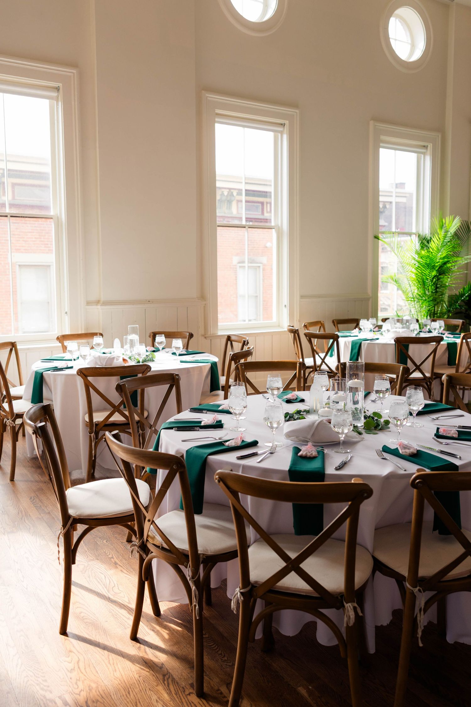 Bright wedding reception hall with round tables, emerald green napkins, white linens, and rustic cross-back wooden chairs.