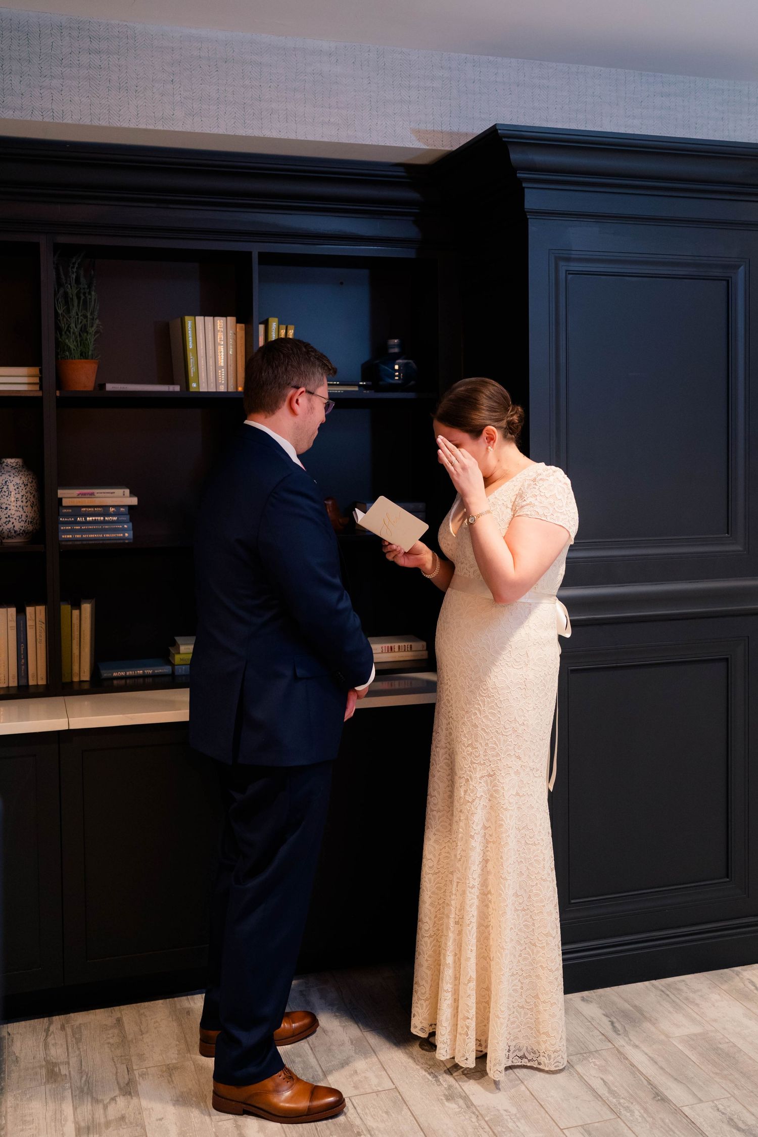 Tearful bride covers face with hand as groom reads moving wedding vows in stylish venue.