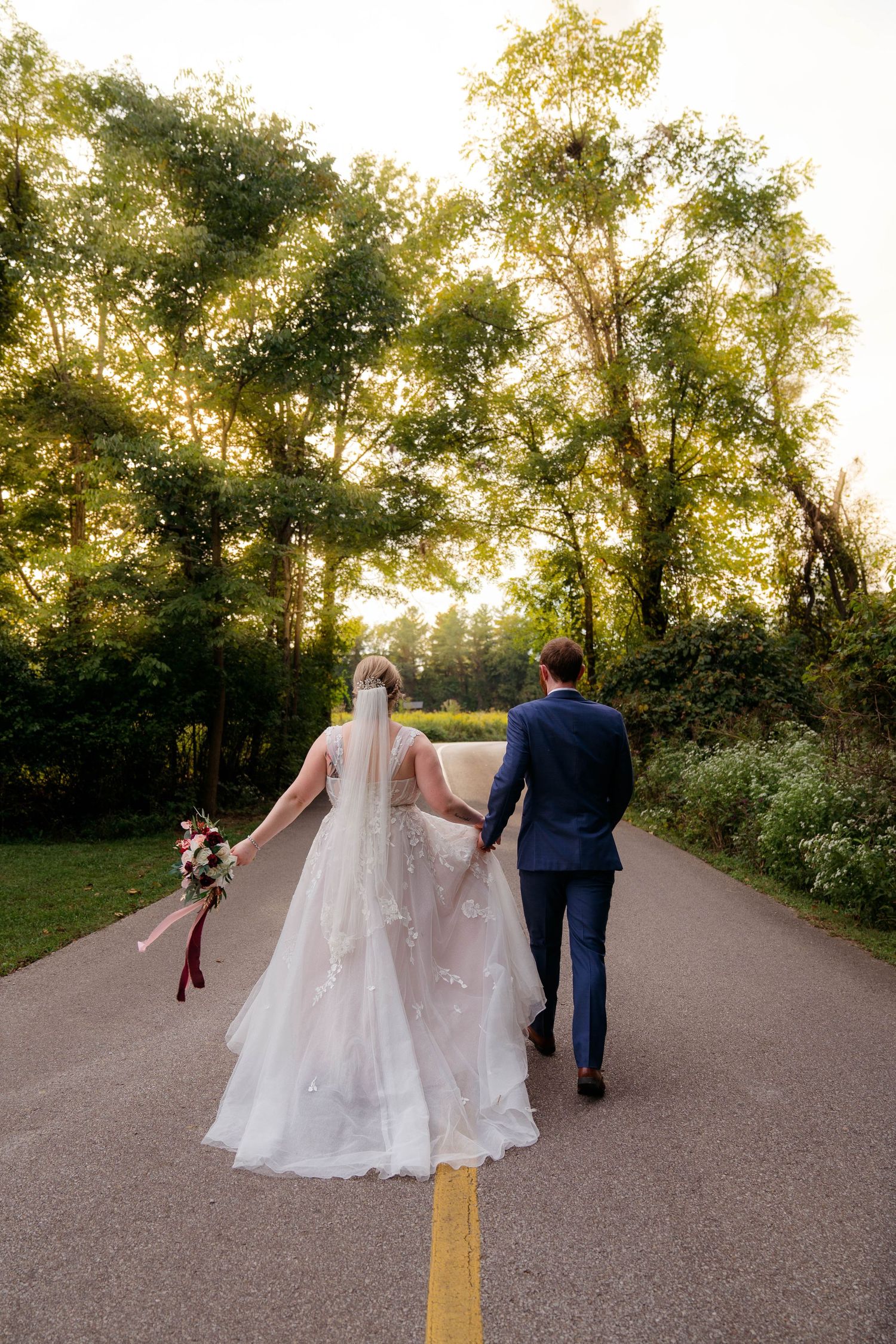 Bride and groom walk hand in hand down a tree-lined road at golden hour sunset.