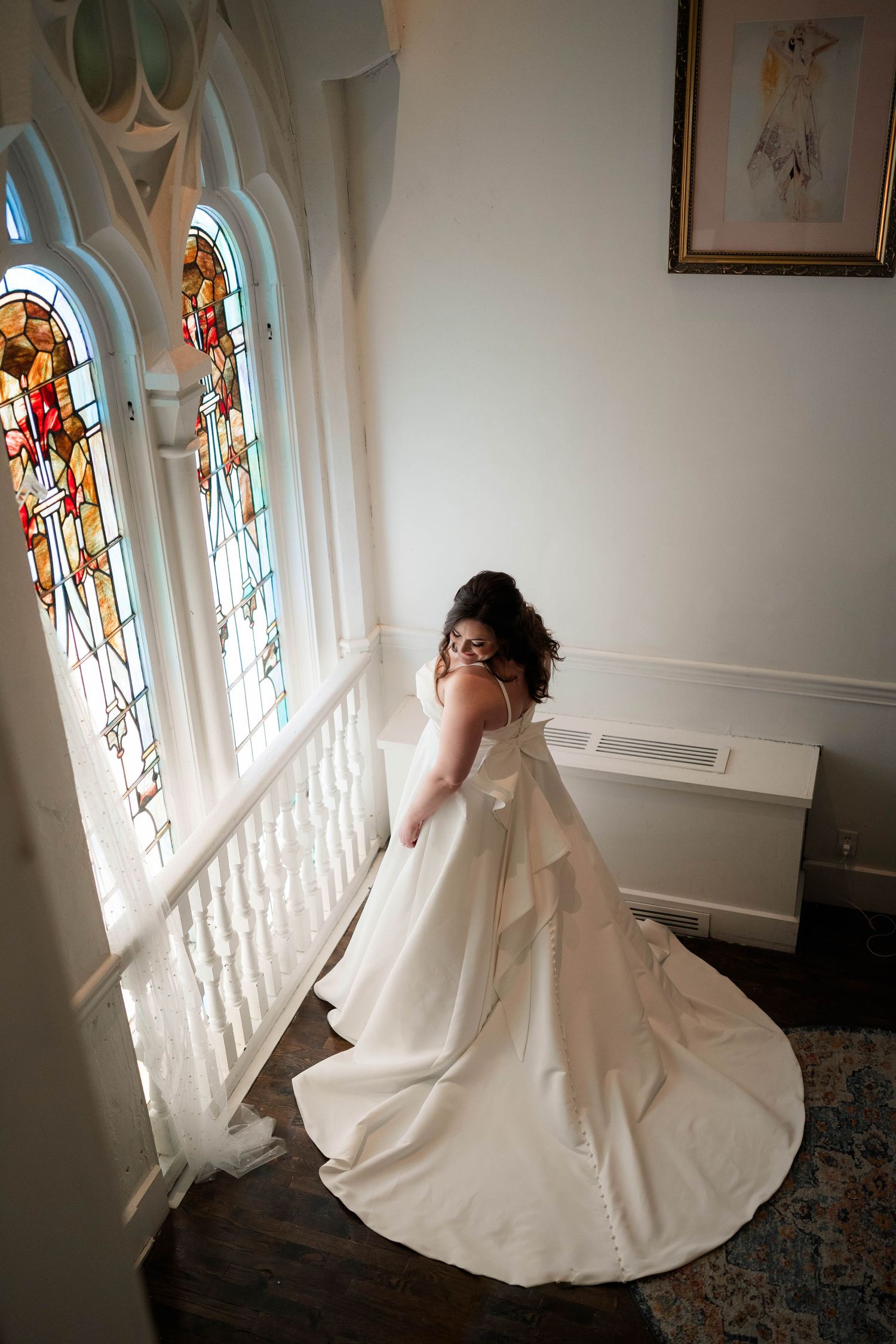Bride in elegant white gown with train stands by stained glass windows on a staircase.