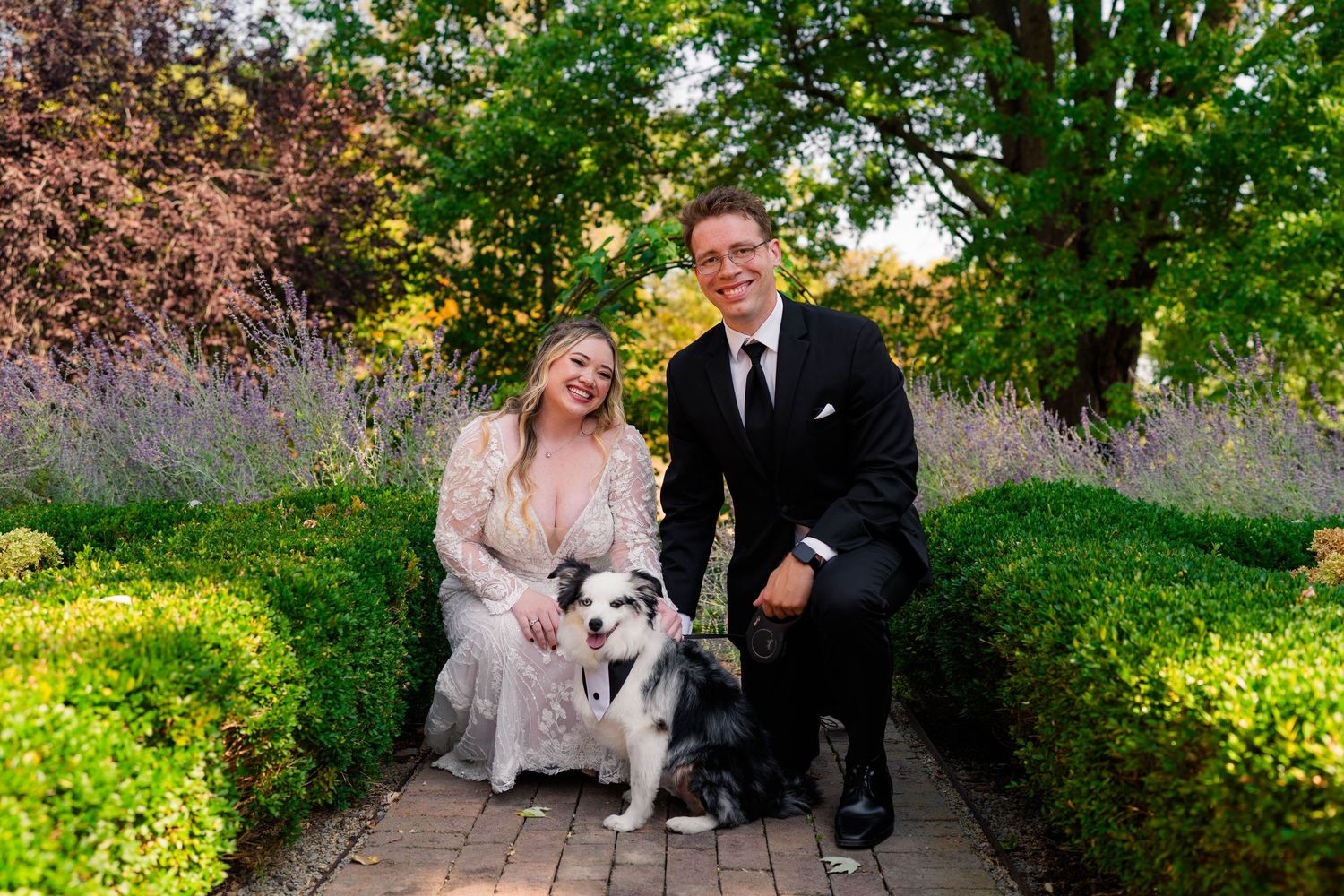 Smiling couple in wedding attire pose with their black and white dog in a lush garden setting.