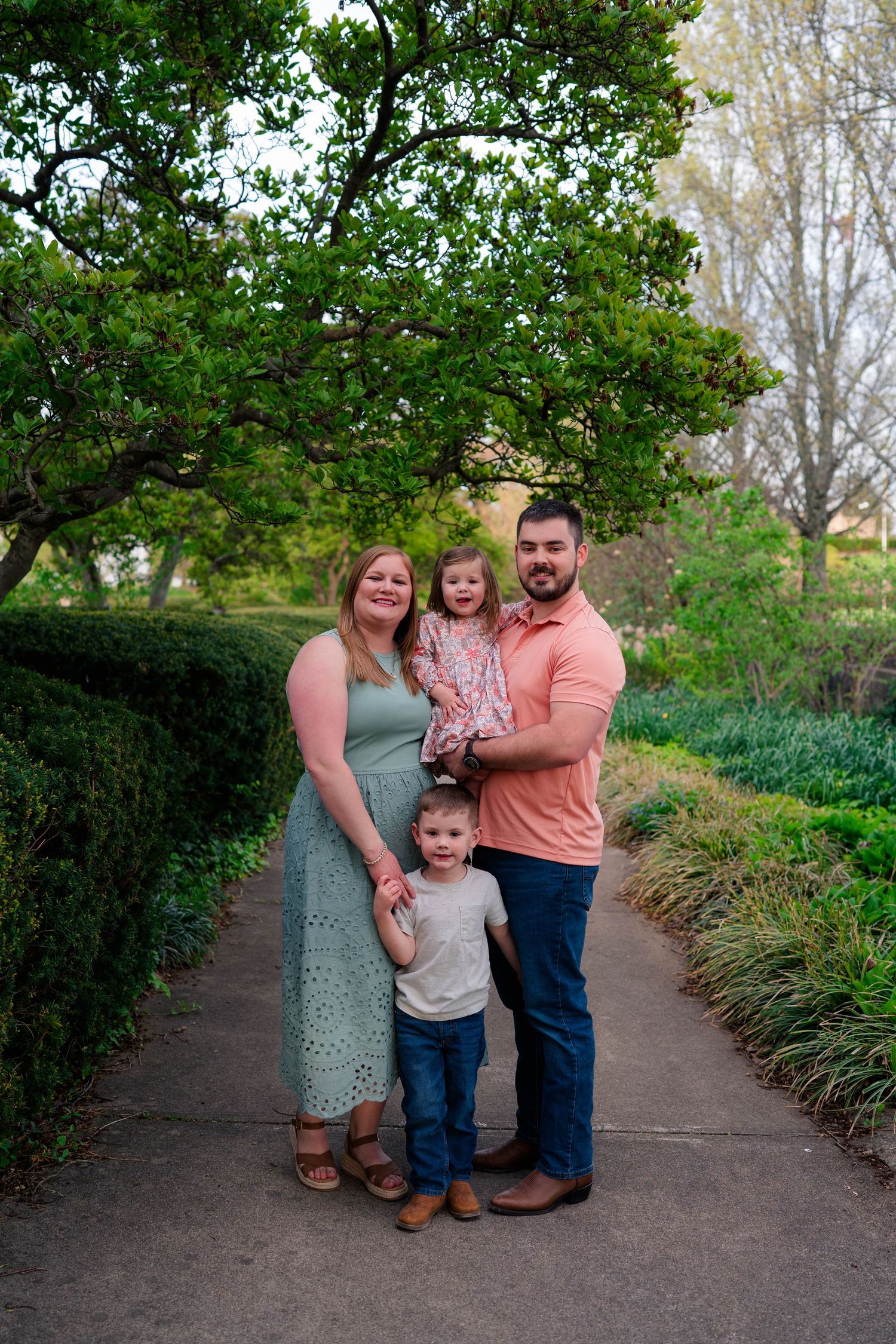 Family of four posing on a garden path surrounded by lush green trees and hedges during a spring photo session.