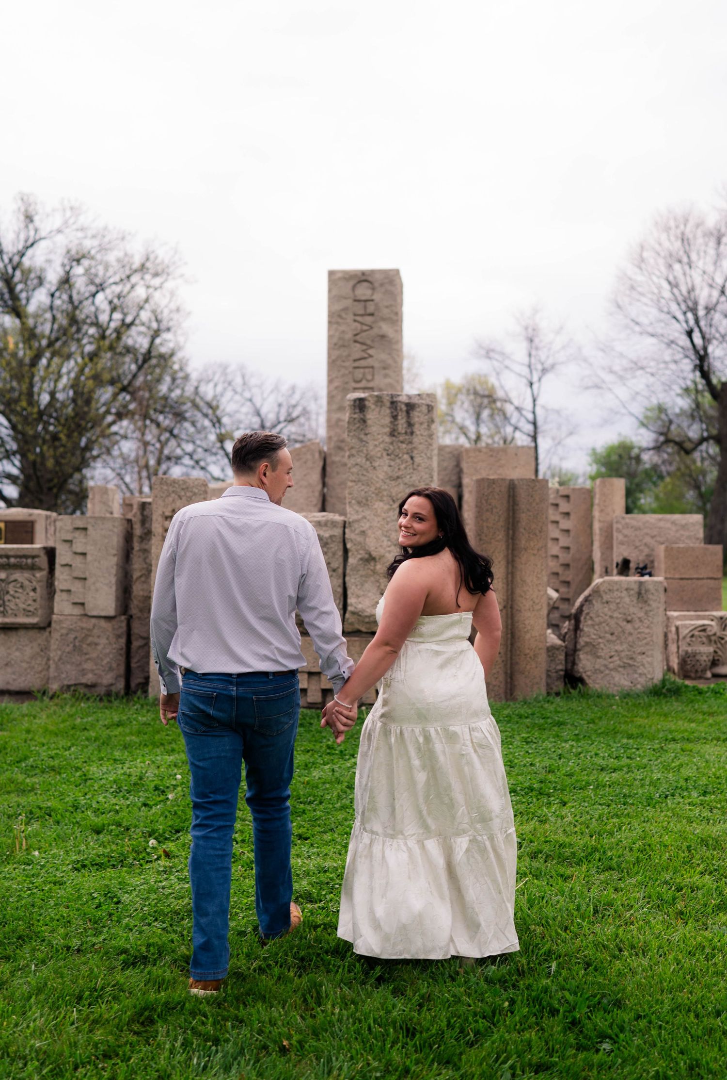 Couple holding hands walking toward stone ruins in a lush green park, woman in white dress.