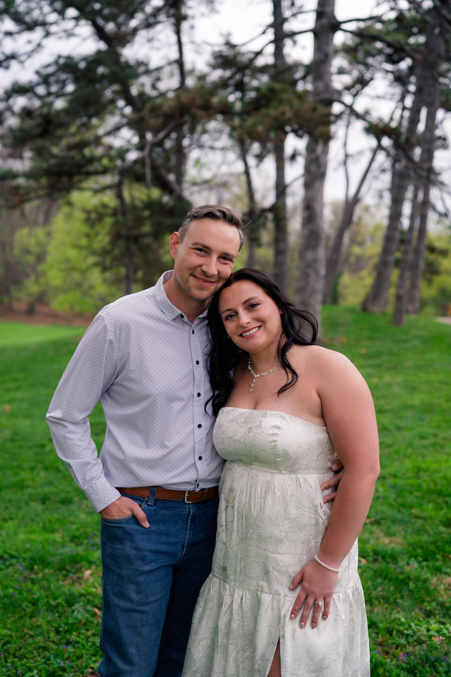 Smiling engaged couple posing together on green lawn surrounded by tall pine trees.