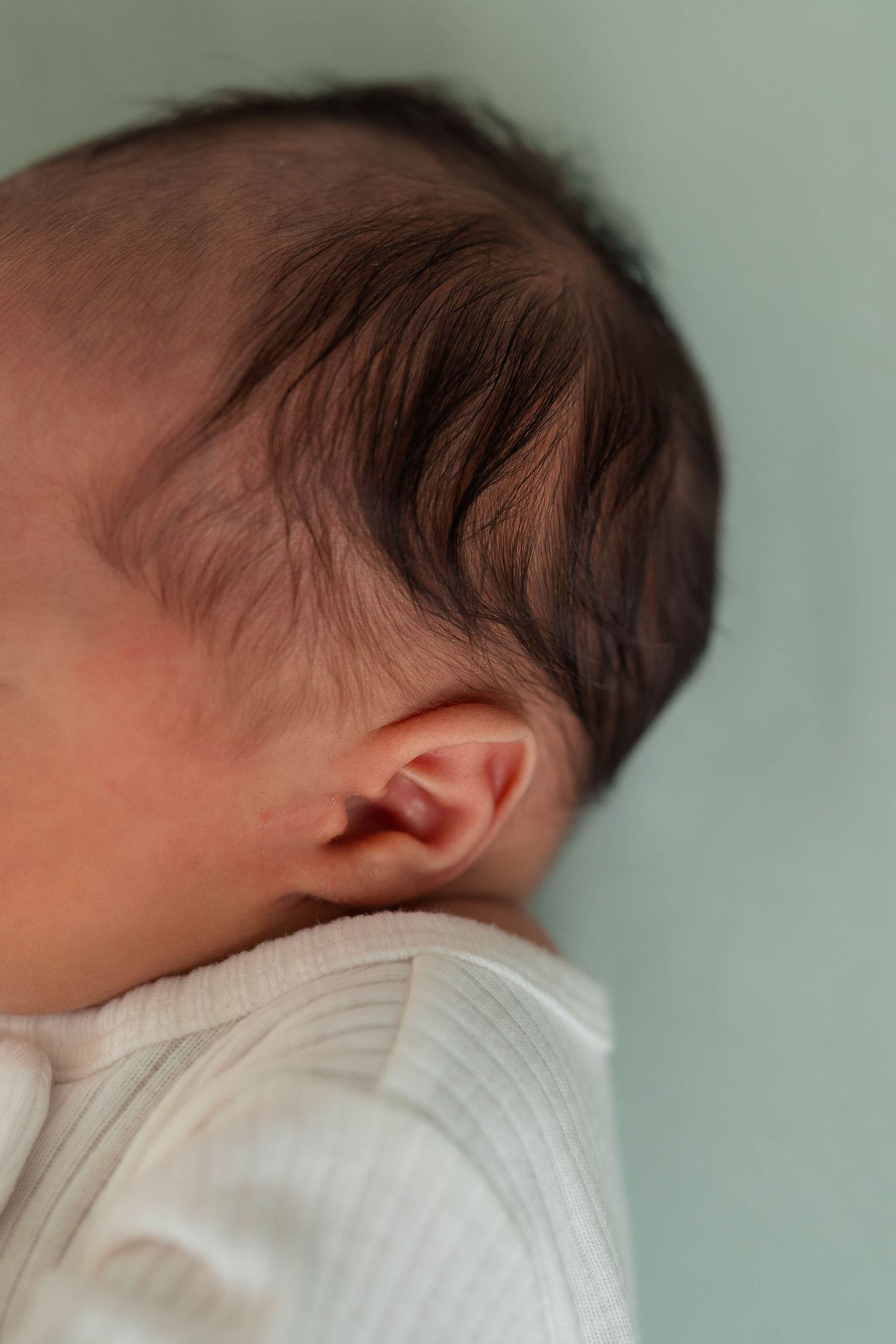 Close-up of newborn baby ear and dark hair in white onesie.