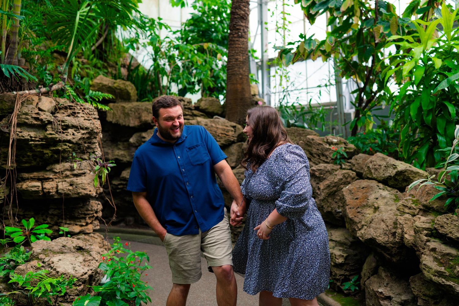 Engaged couple holding hands and walking together along a path through a lush tropical garden.