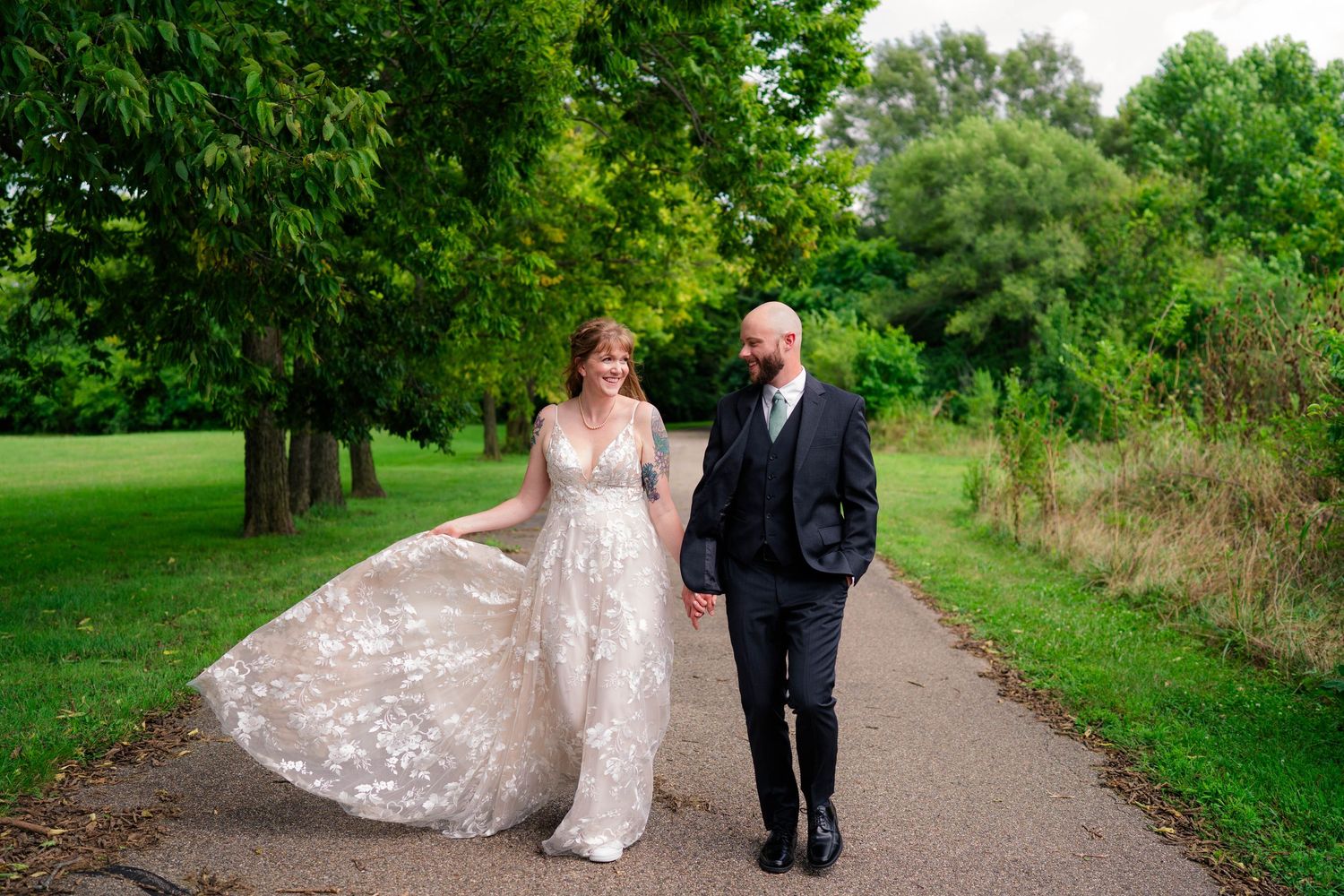 Bride and groom walk hand in hand on a garden path, bride wearing a floral lace gown as her dress flows in the breeze.