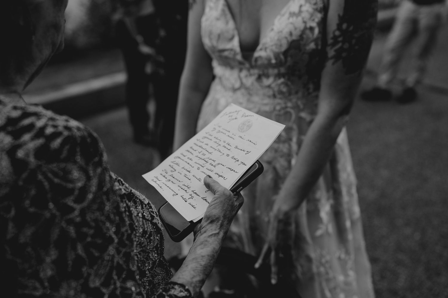 Black and white photo of an elderly guest reading a handwritten card near a bride in a lace gown.