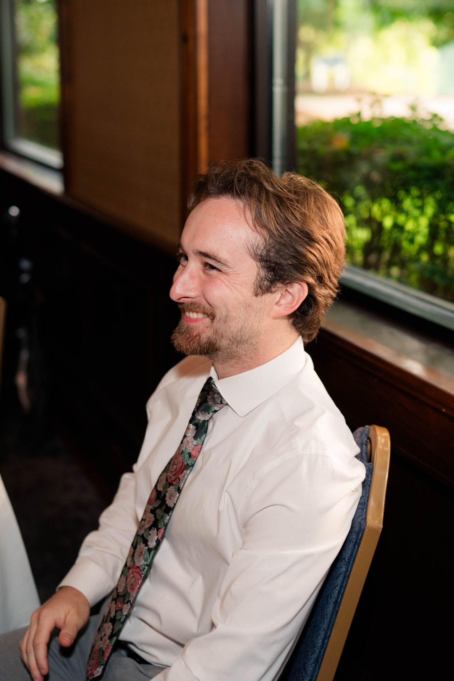 Smiling man with floral tie sits by window at indoor wedding reception celebration.