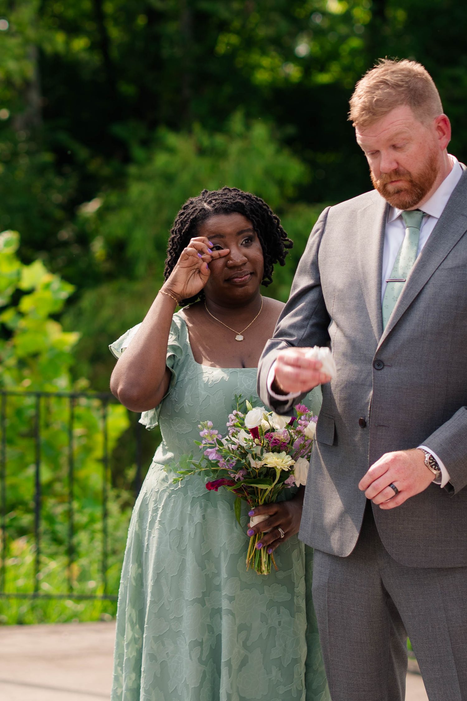 Emotional bridesmaid in a sage green dress wipes tears while standing beside a red-haired groomsman holding her hand.