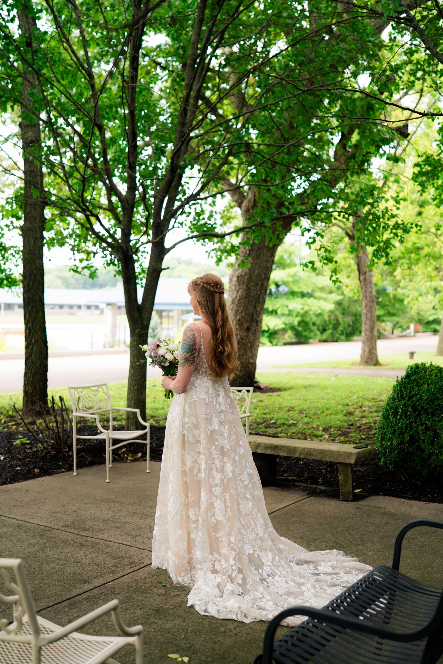 A bride in a lace wedding gown with a train holds a bouquet while standing under green trees outdoors.