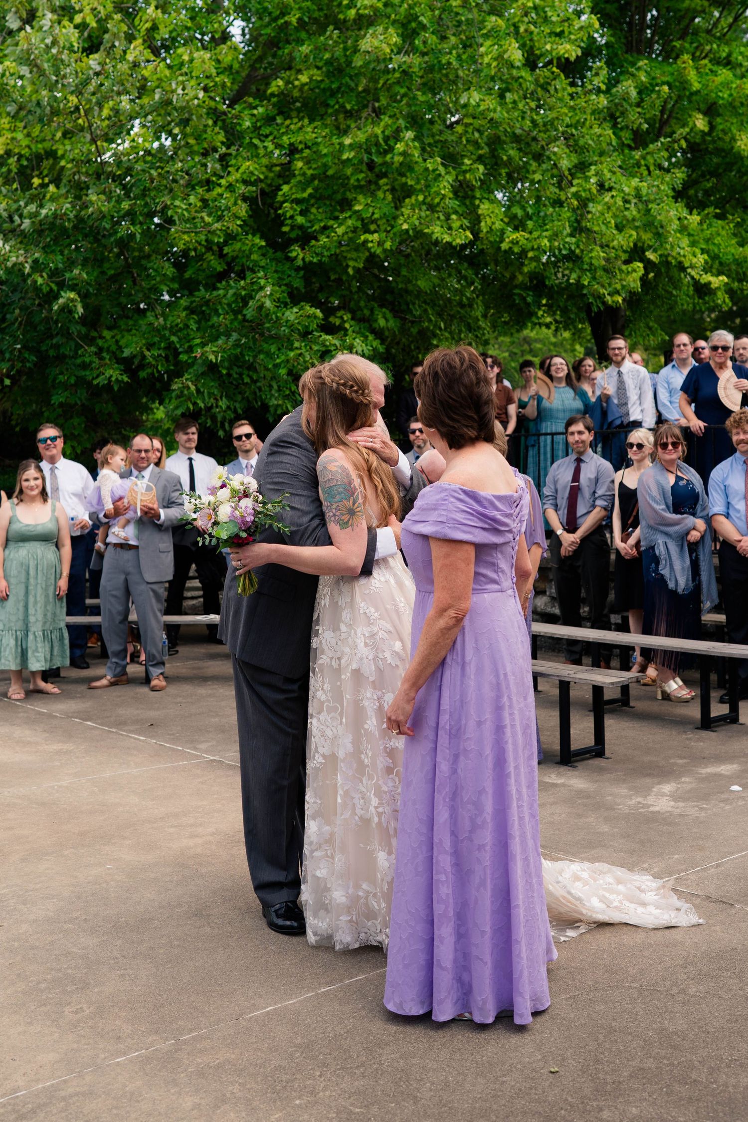 Bride and groom share a kiss during outdoor wedding ceremony while guests watch and woman in lavender gown stands nearby.