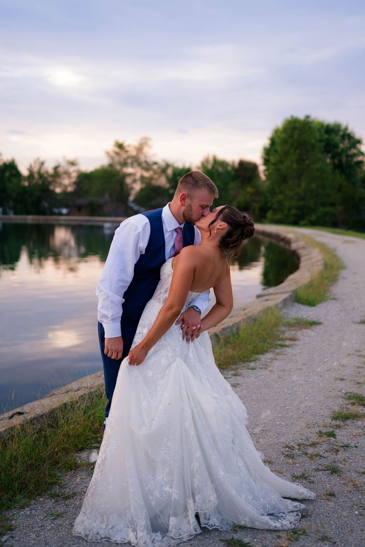 Bride and groom share a romantic kiss by a serene lake at sunset in wedding attire.