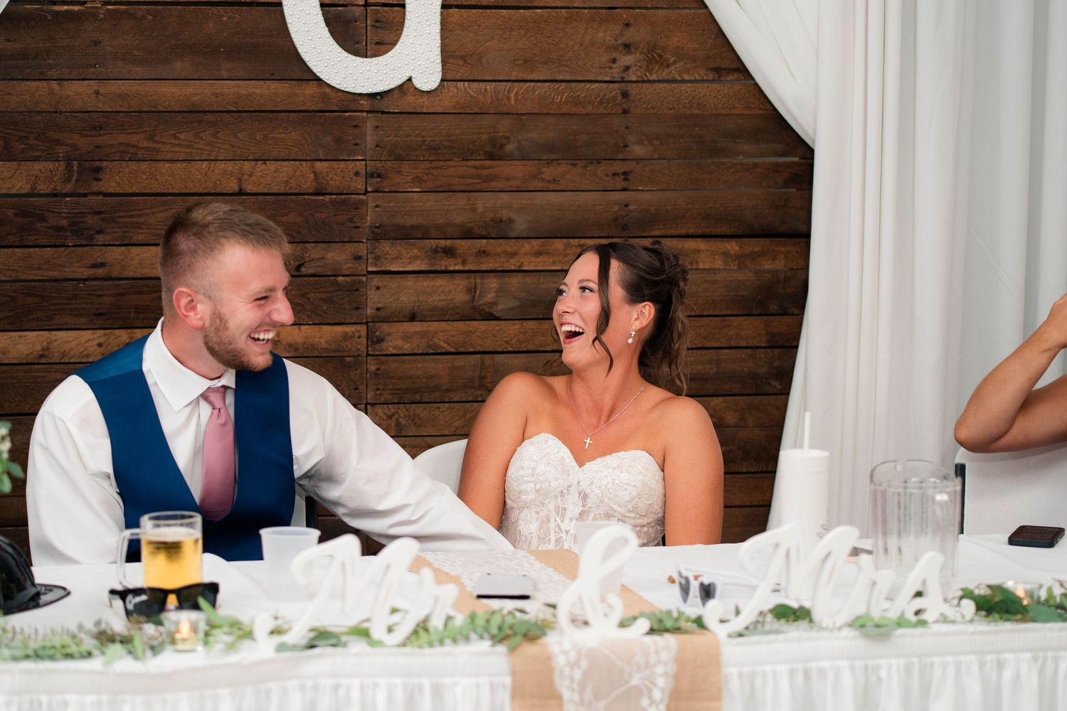 Bride and groom laughing joyfully at wedding reception head table with rustic wood backdrop.