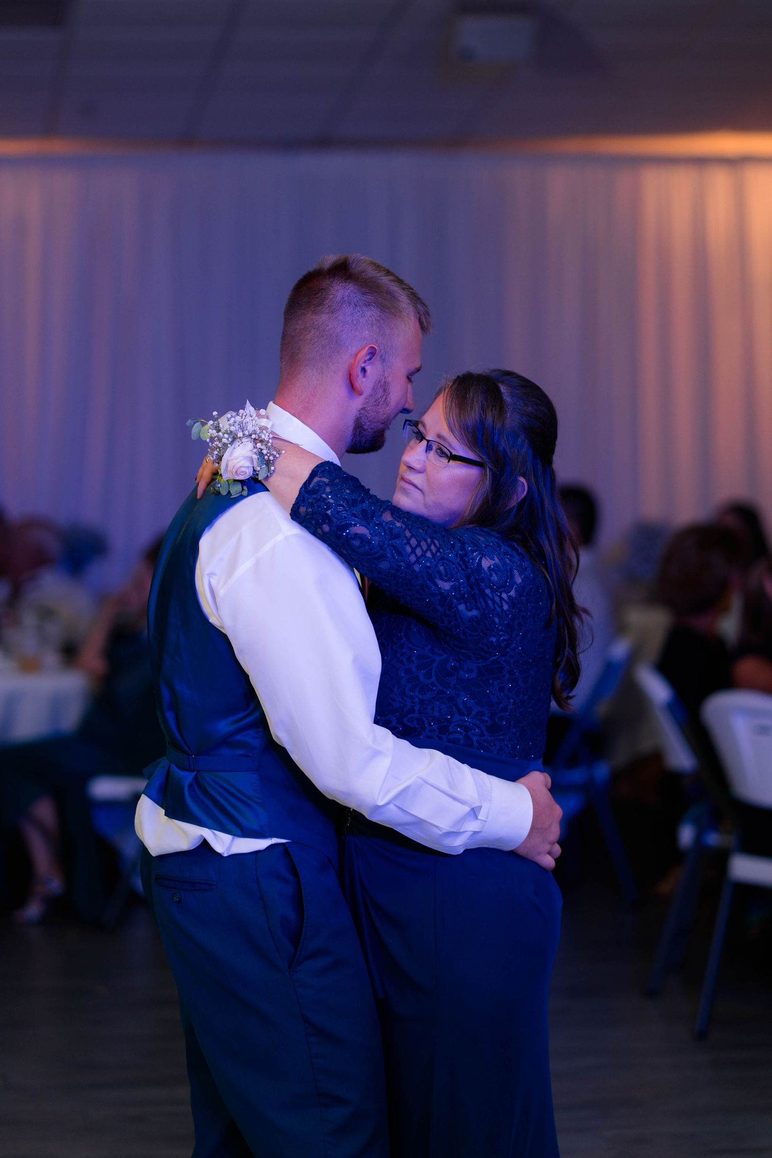 Groom shares emotional mother-son dance under blue lighting at elegant wedding reception venue.